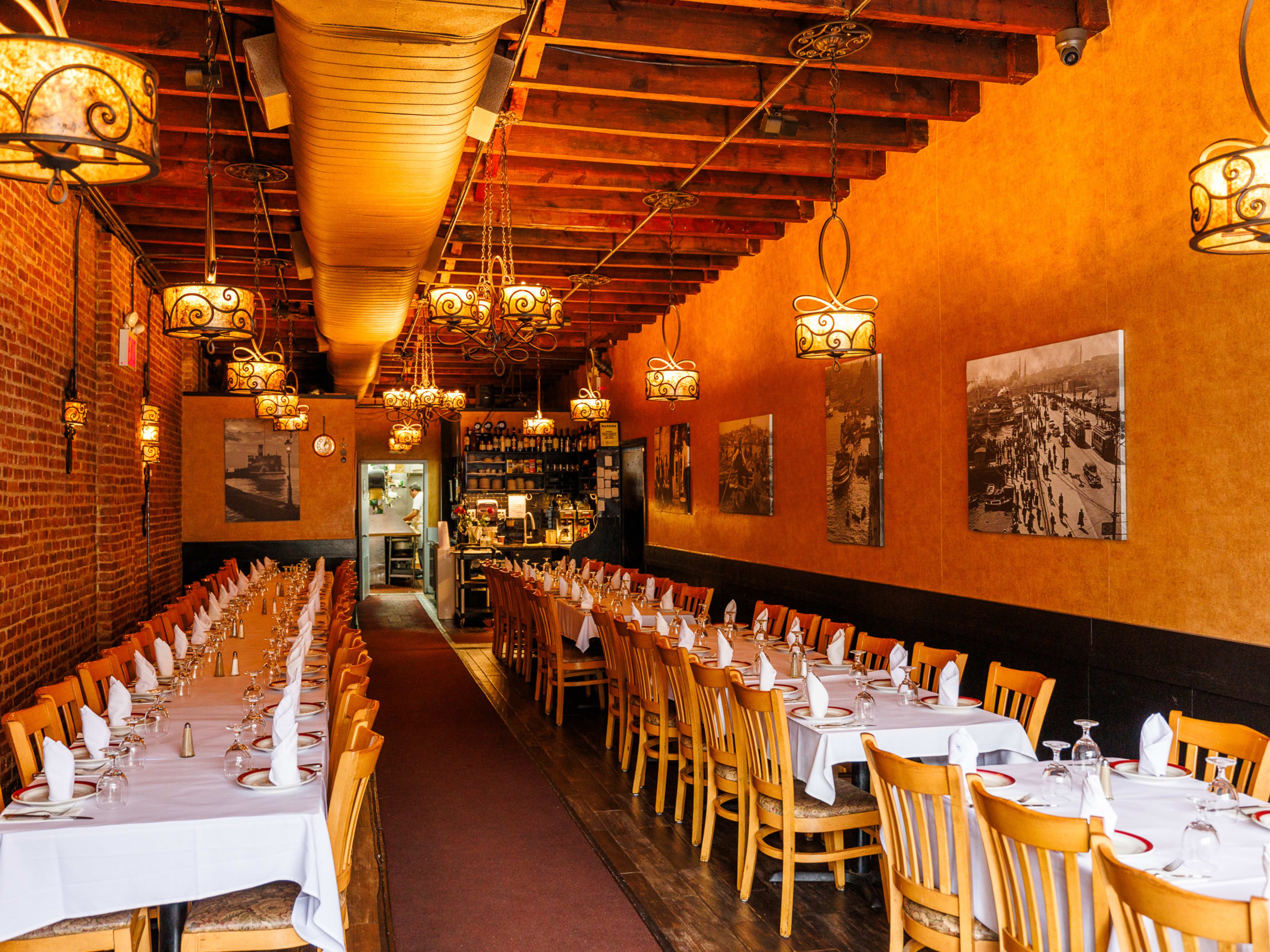 Taci's Beyti Restaurant interiors with orange walls, warm orange light fixtures hanging from the ceiling, and two long rows of square dining tables with white tablecloths and place settings