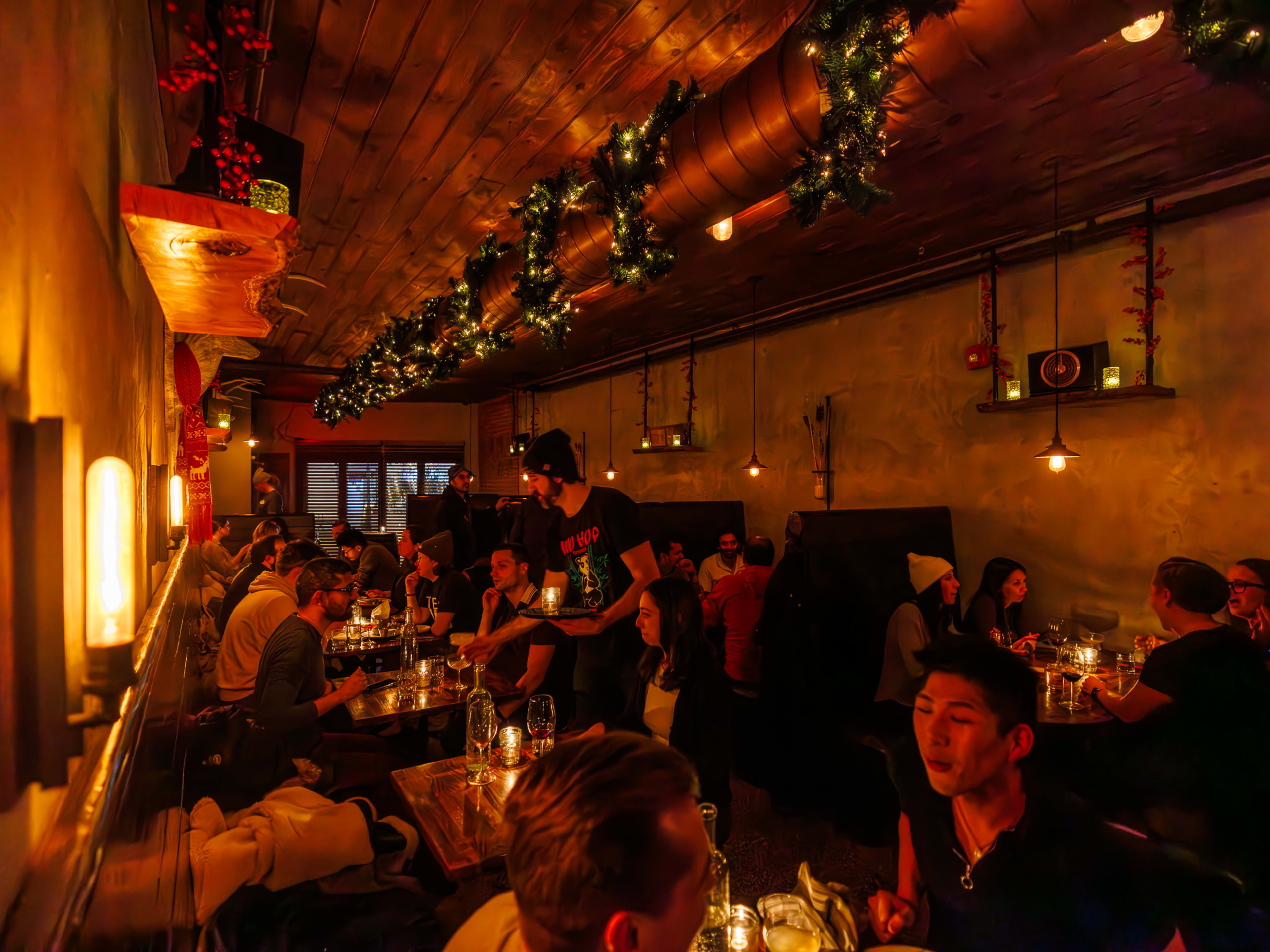 The Archer interiors at night with very dim lighting, people seated at wooden tables with candles on them, and garland with twinkle lights twisted around a pipe on the ceiling.