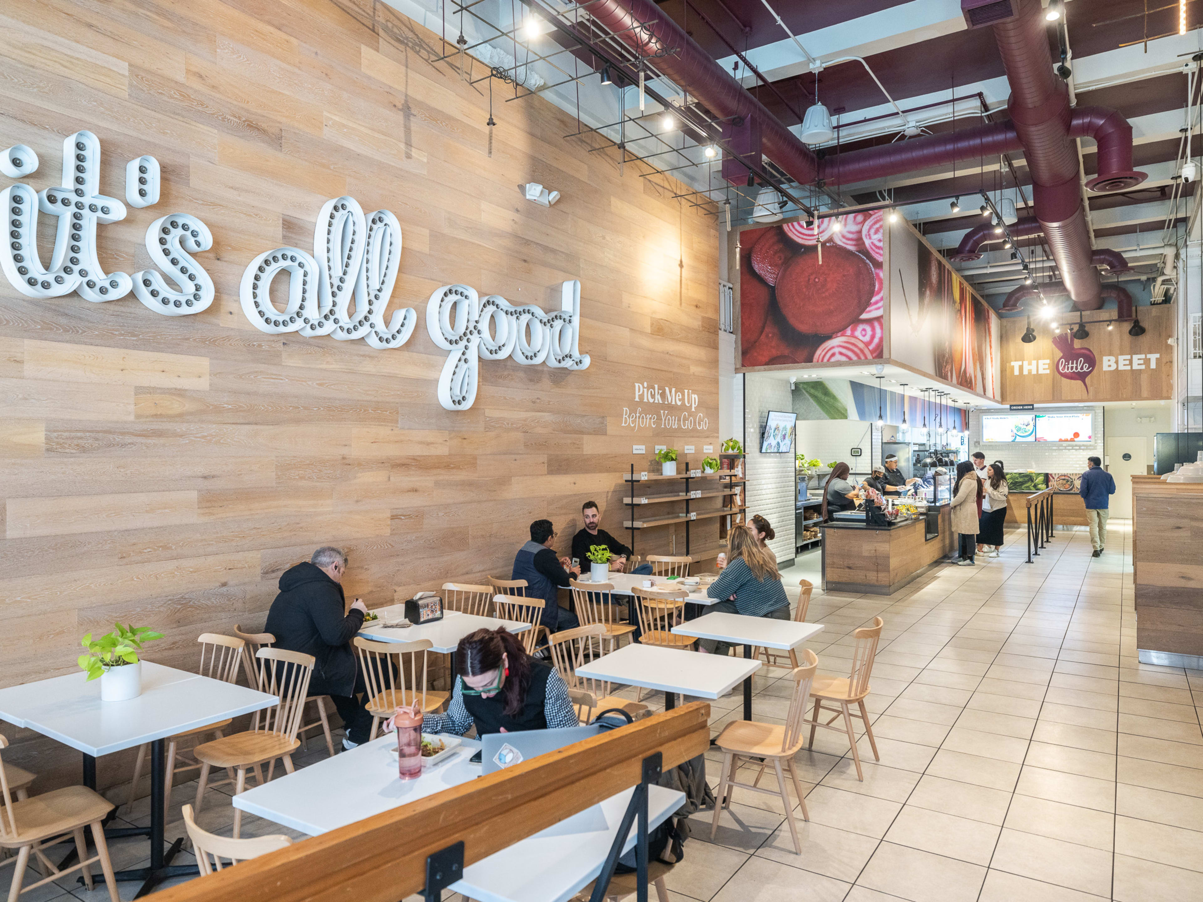 The Little Beet interiors with light wood walls, a large sign on the left wall, tile floors, and an assembly line counter in the back with customers placing orders