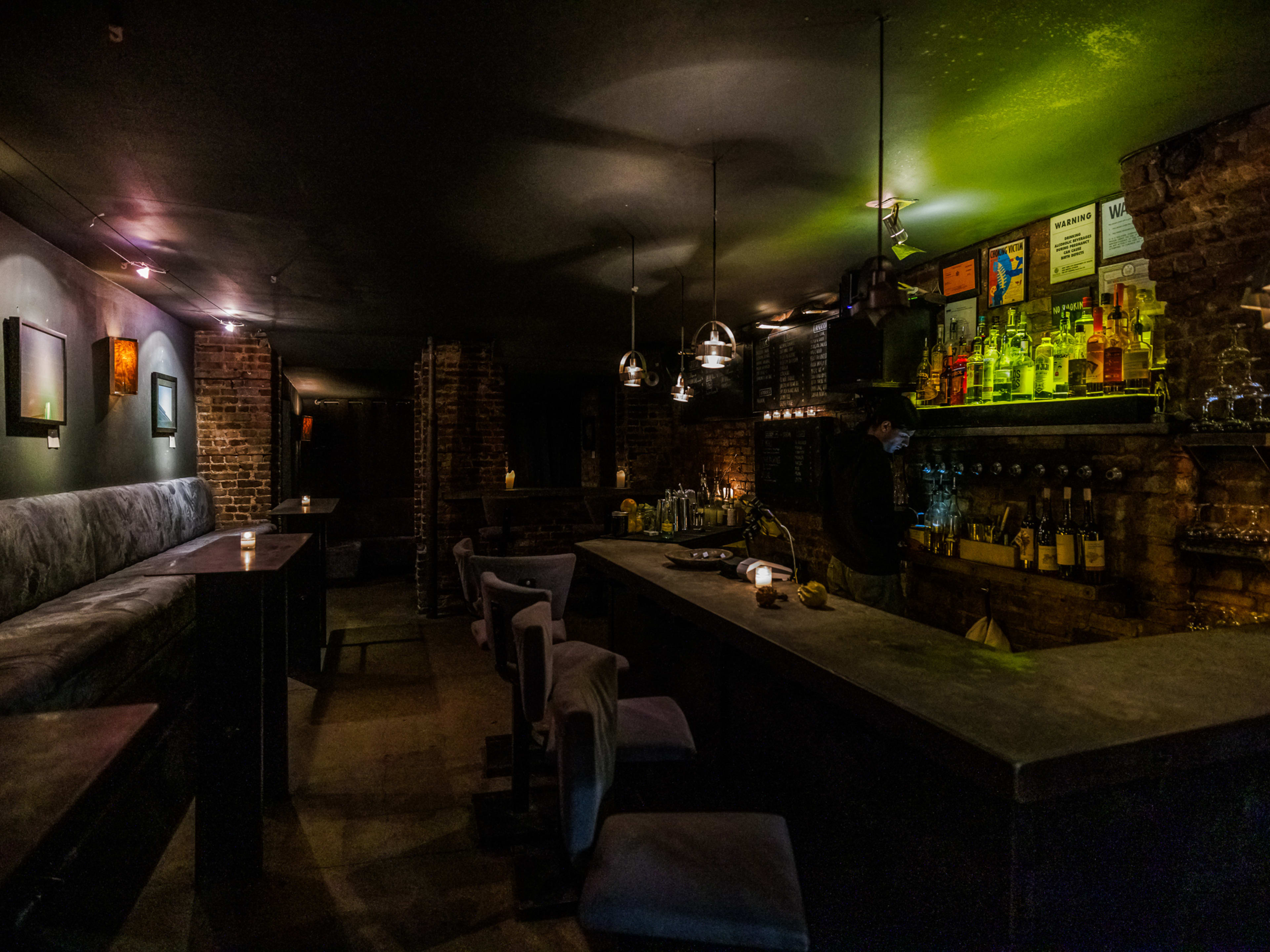 The Otheroom interiors with very dim lighting, candles on tables with booth seating against the left wall, and cushioned stools at a stone bar to the right