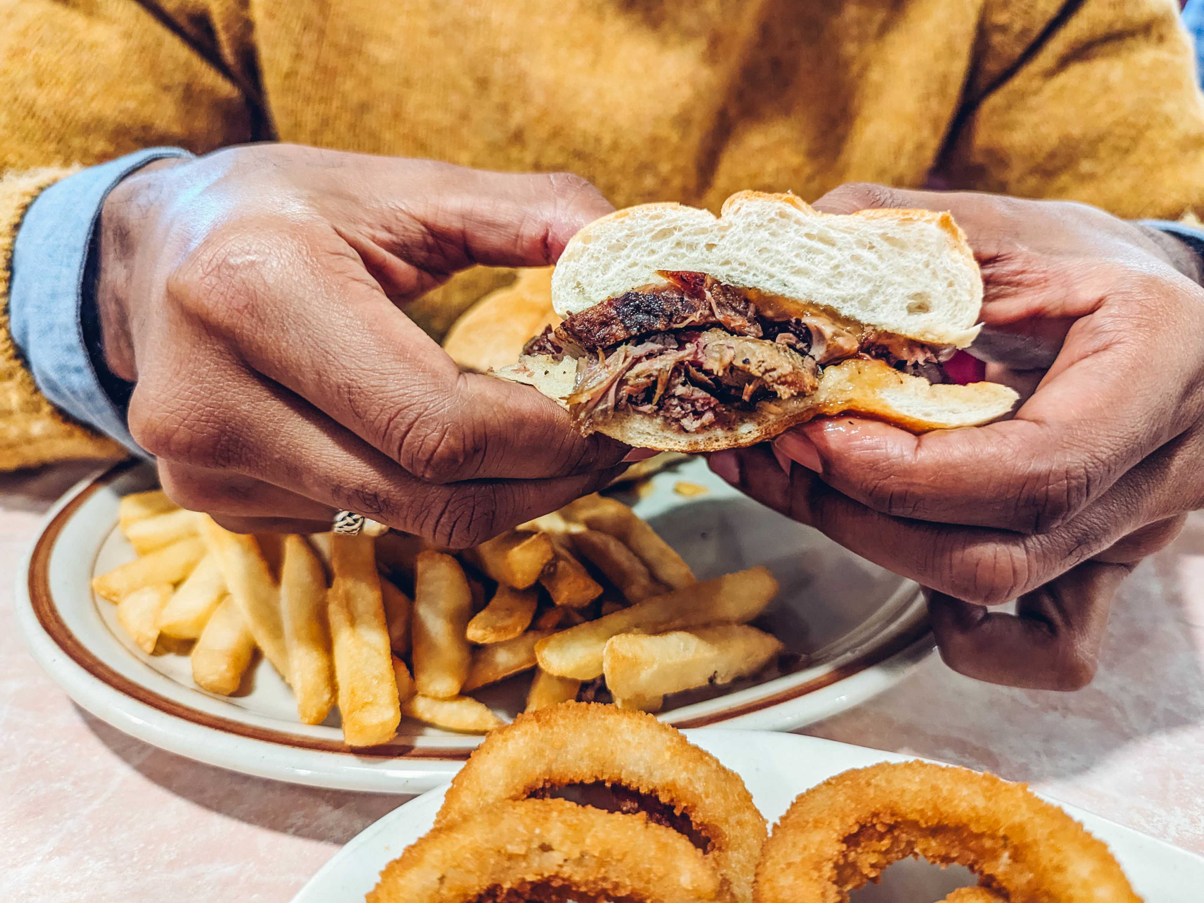 hands holding a roast leg of lamb sandwich, with fries and onion rings