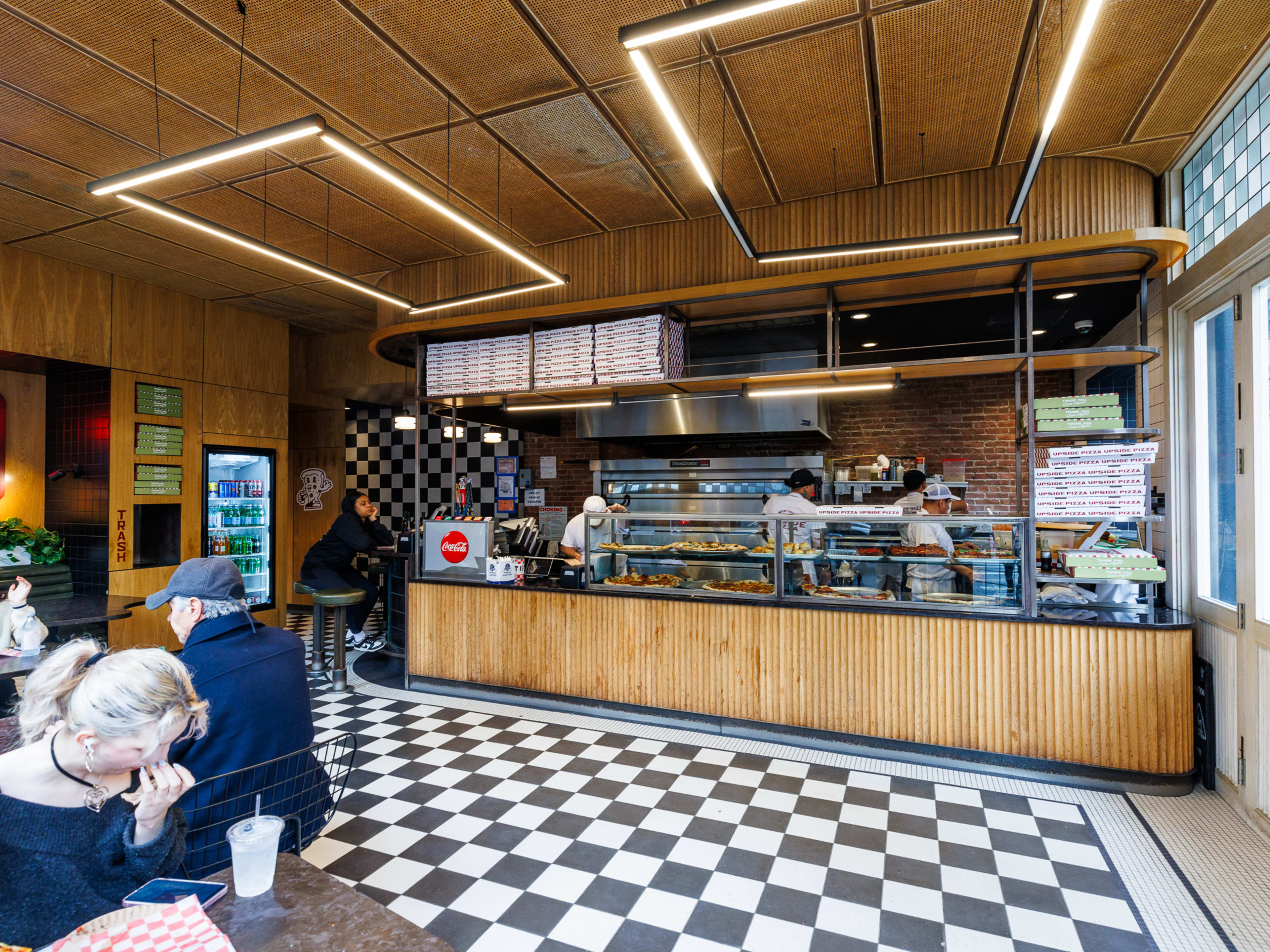 Upside Pizza interiors with black and white checkerboard tile floors, a large counter with pizzas on display behind glass, and customers seated at tables to the left