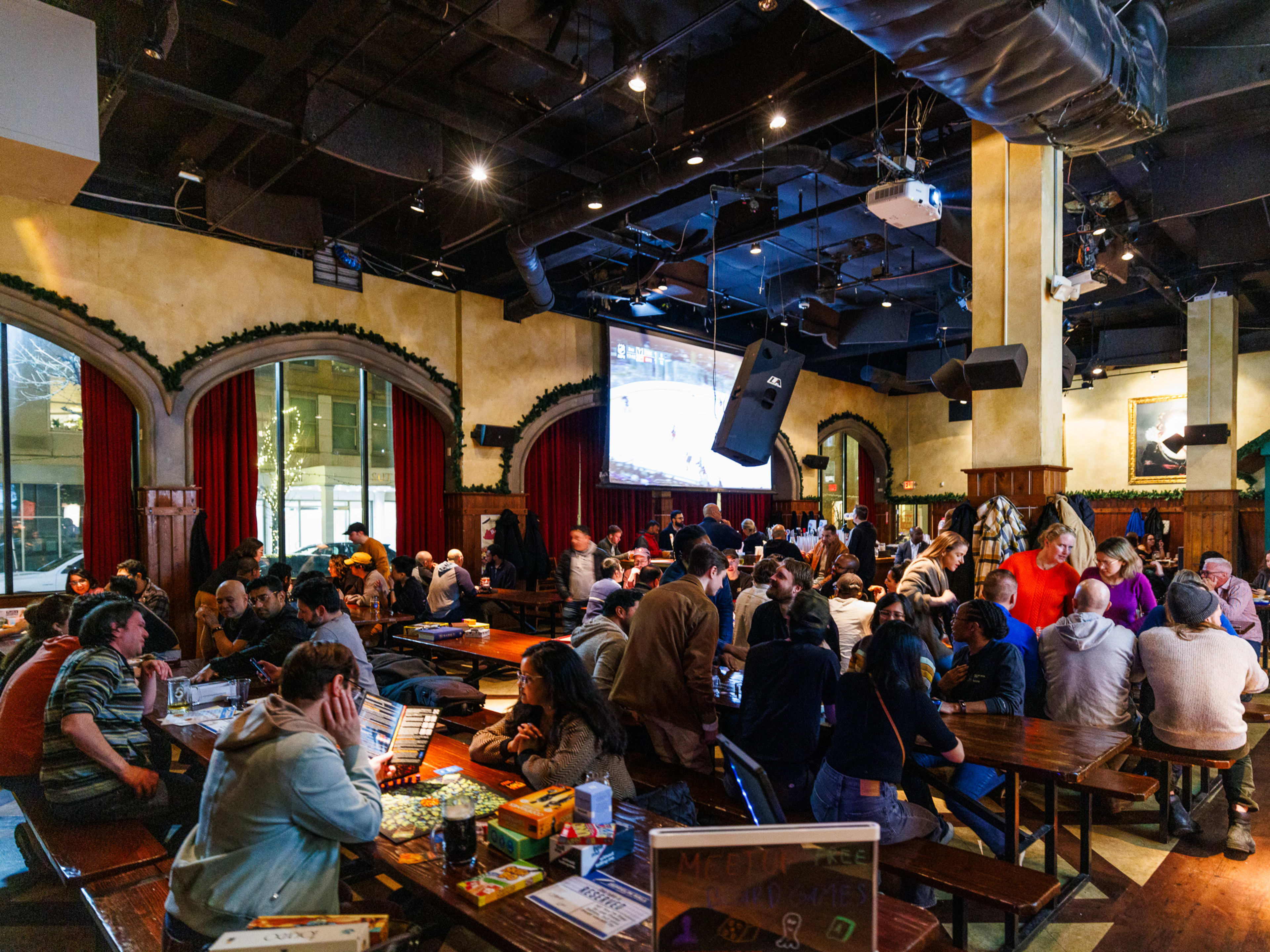 Zeppelin Hall Biergarten interiors with groups of people seated at wooden picnic tables and a large projector screen hanging on the wall