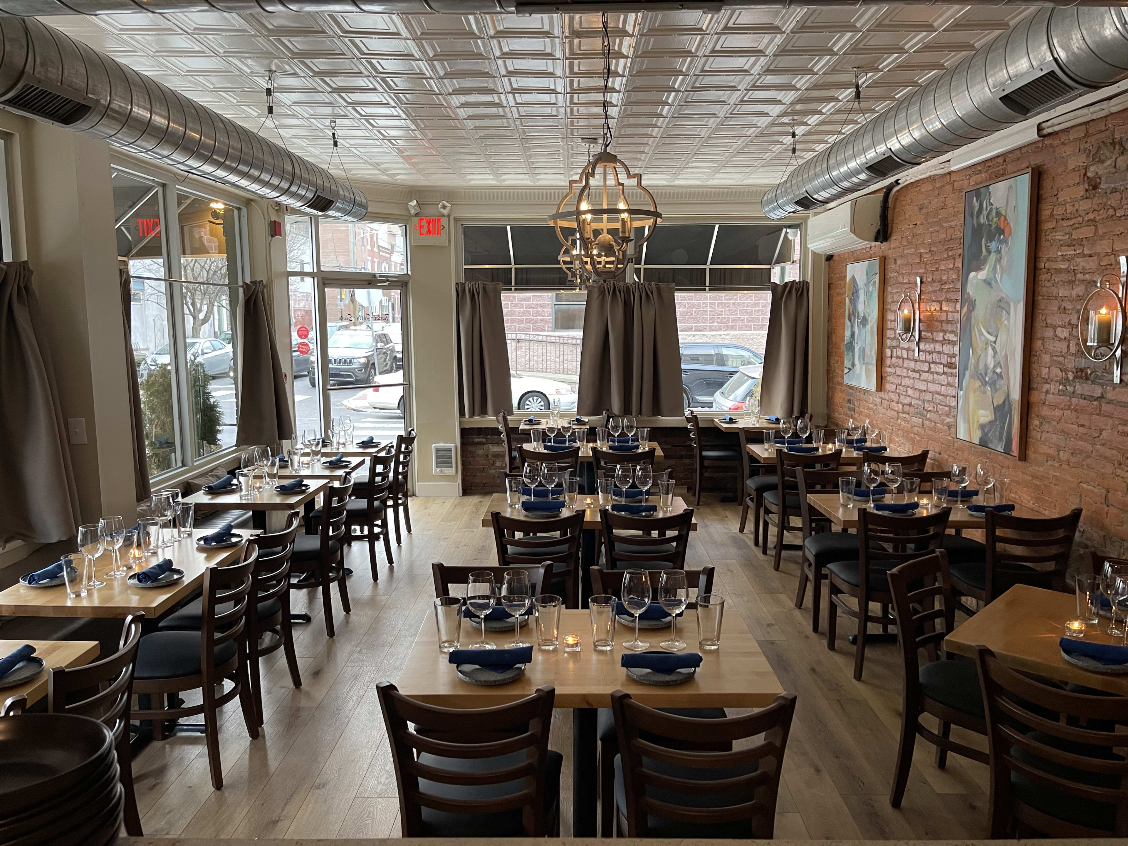 Burrata interiors with brick walls, large windows with a view to the street corner, and rectangular wooden tables all set with place settings and candles