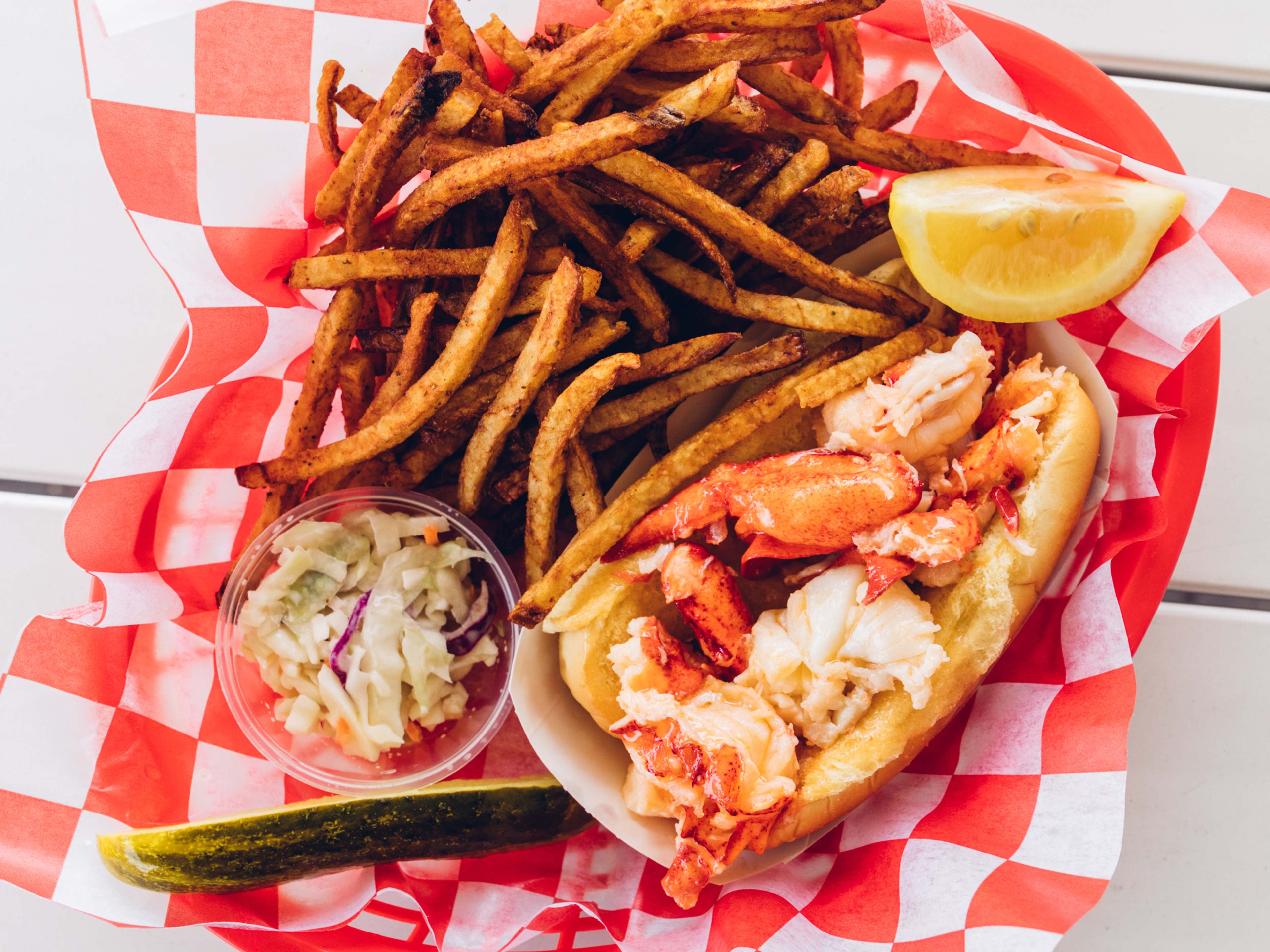 Lobster roll with fries cole slaw and a pickle shot on an outdoor picnic table