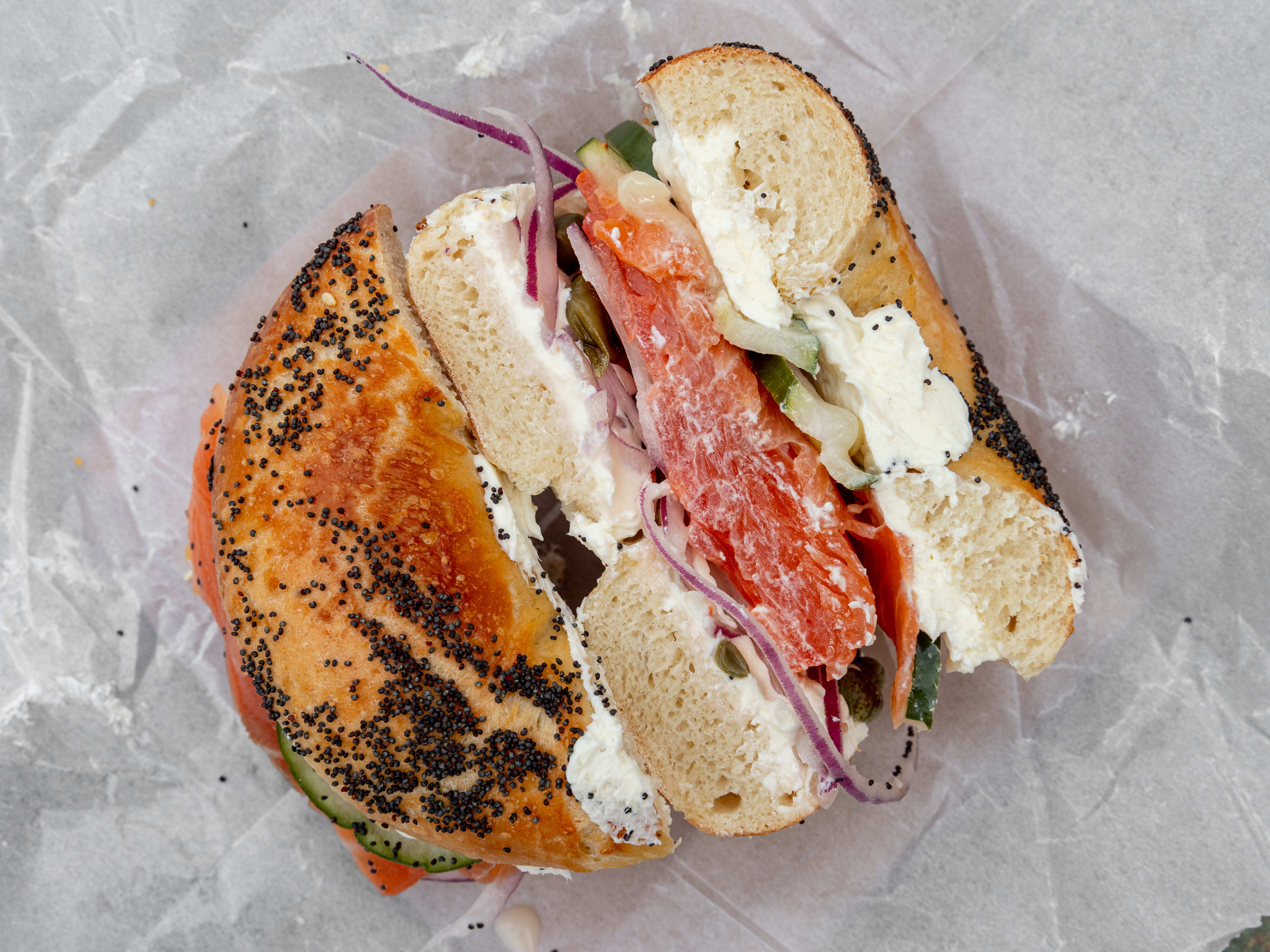 Cross-section of a lox sandwich with layers of cream cheese, cucumber, tomatoes, and onions on a poppy bagel