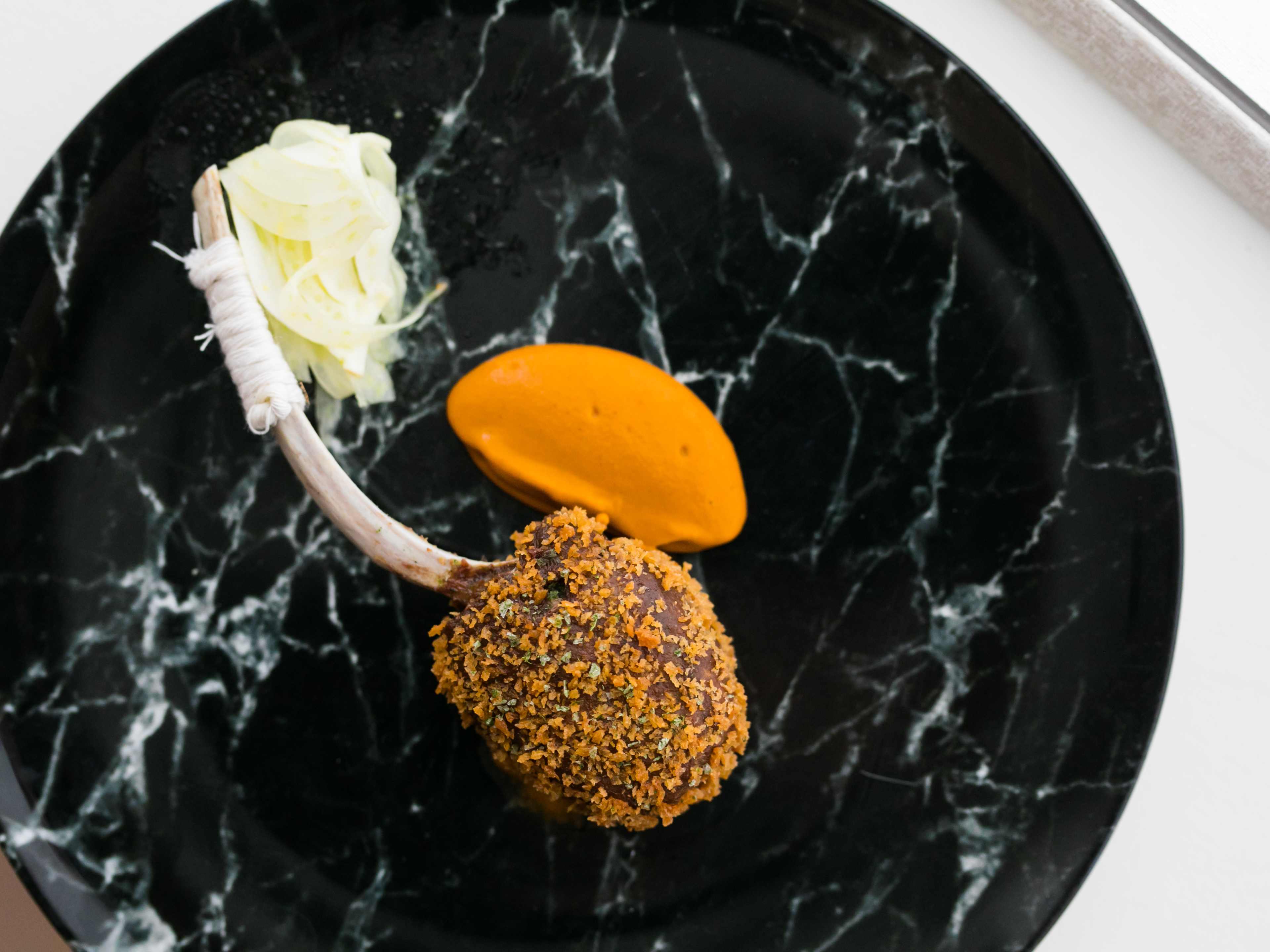 Black marble plate with artfully plated food that looks like orange sorbet and a lamb chop