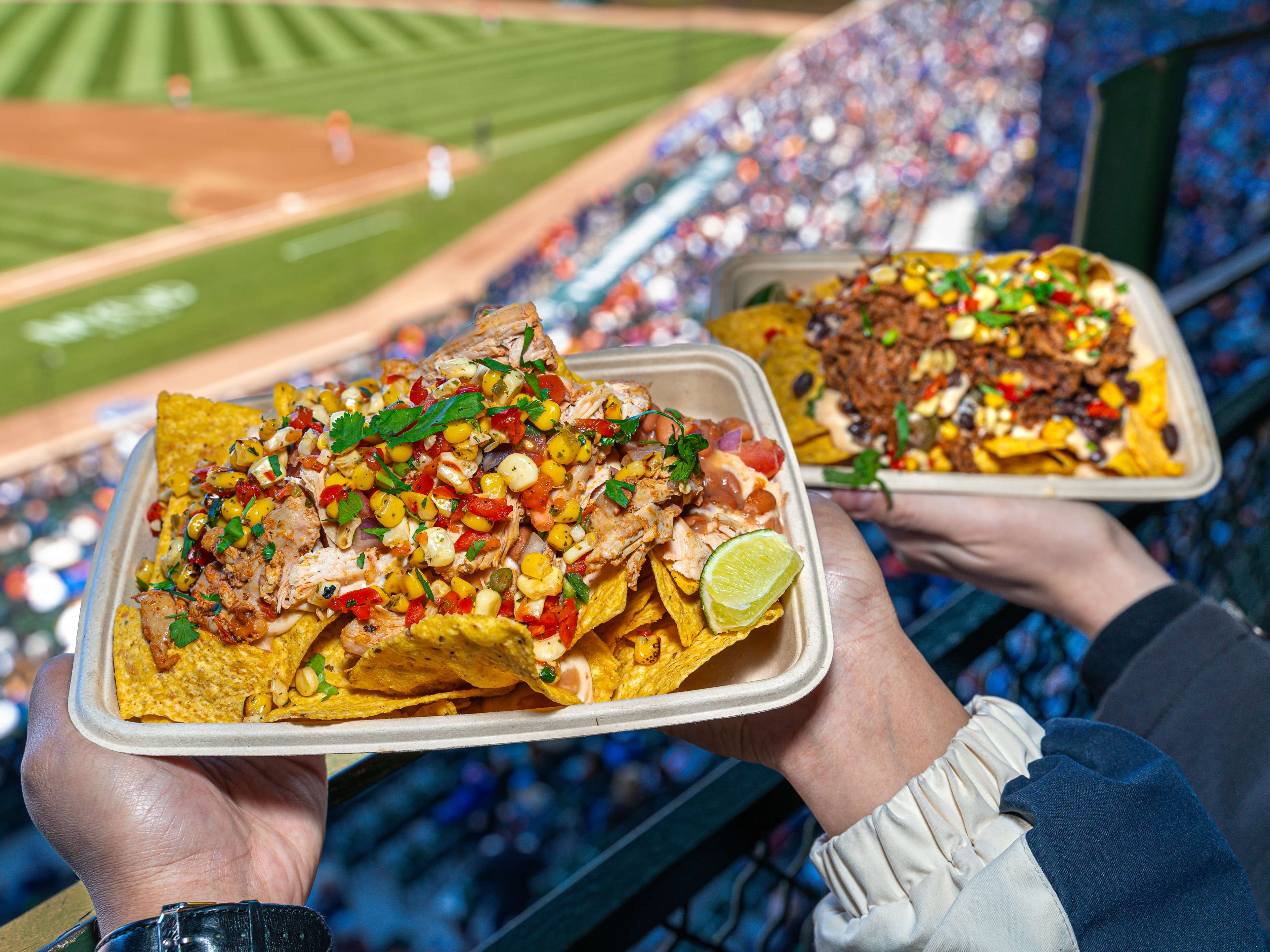 Two hands holding up two plates of loaded nachos at a Cubs game.