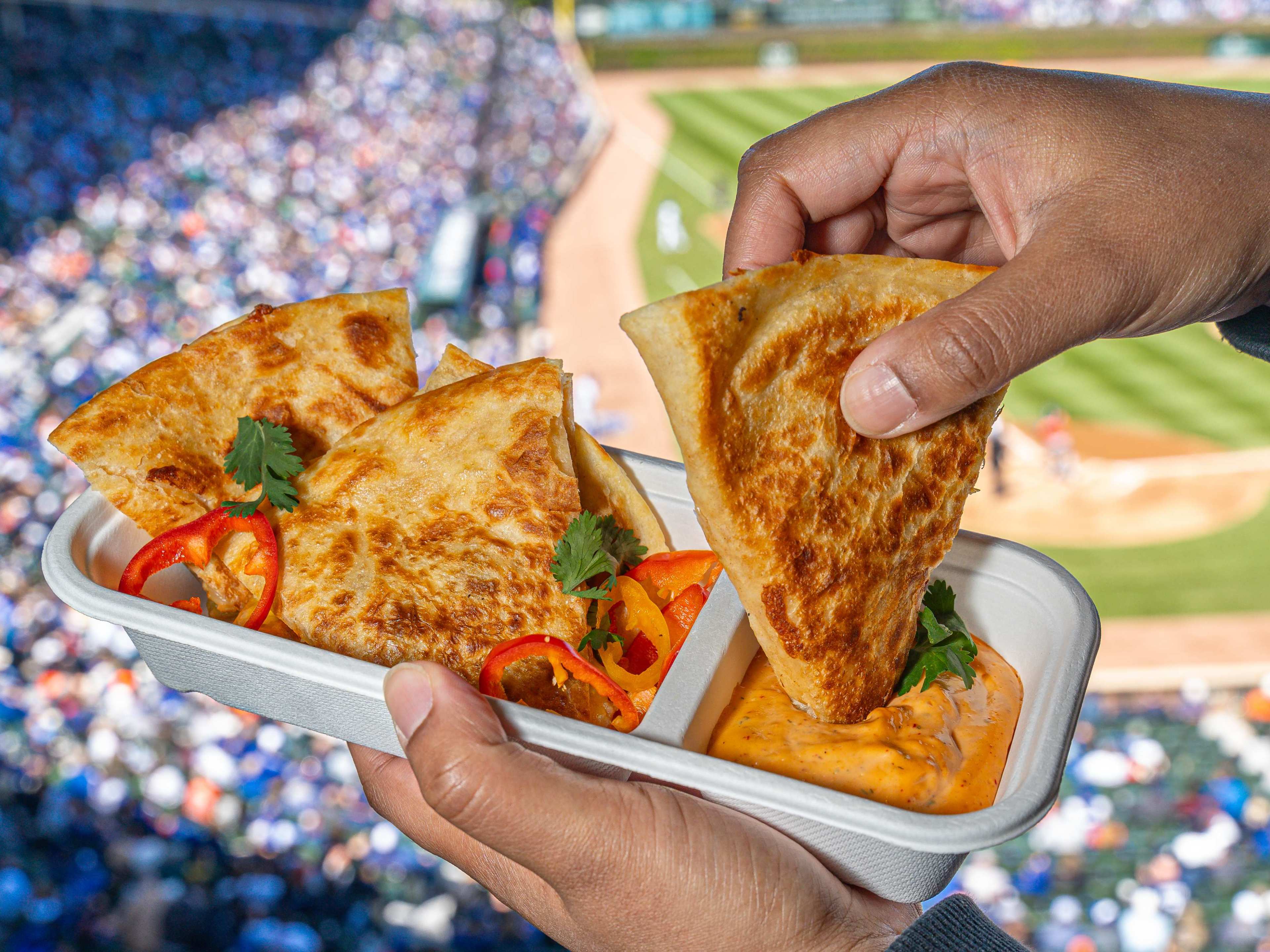 A hand dipping a piece of chicken quesadilla into a chipotle ranch sauce with a Cubs game in the background.