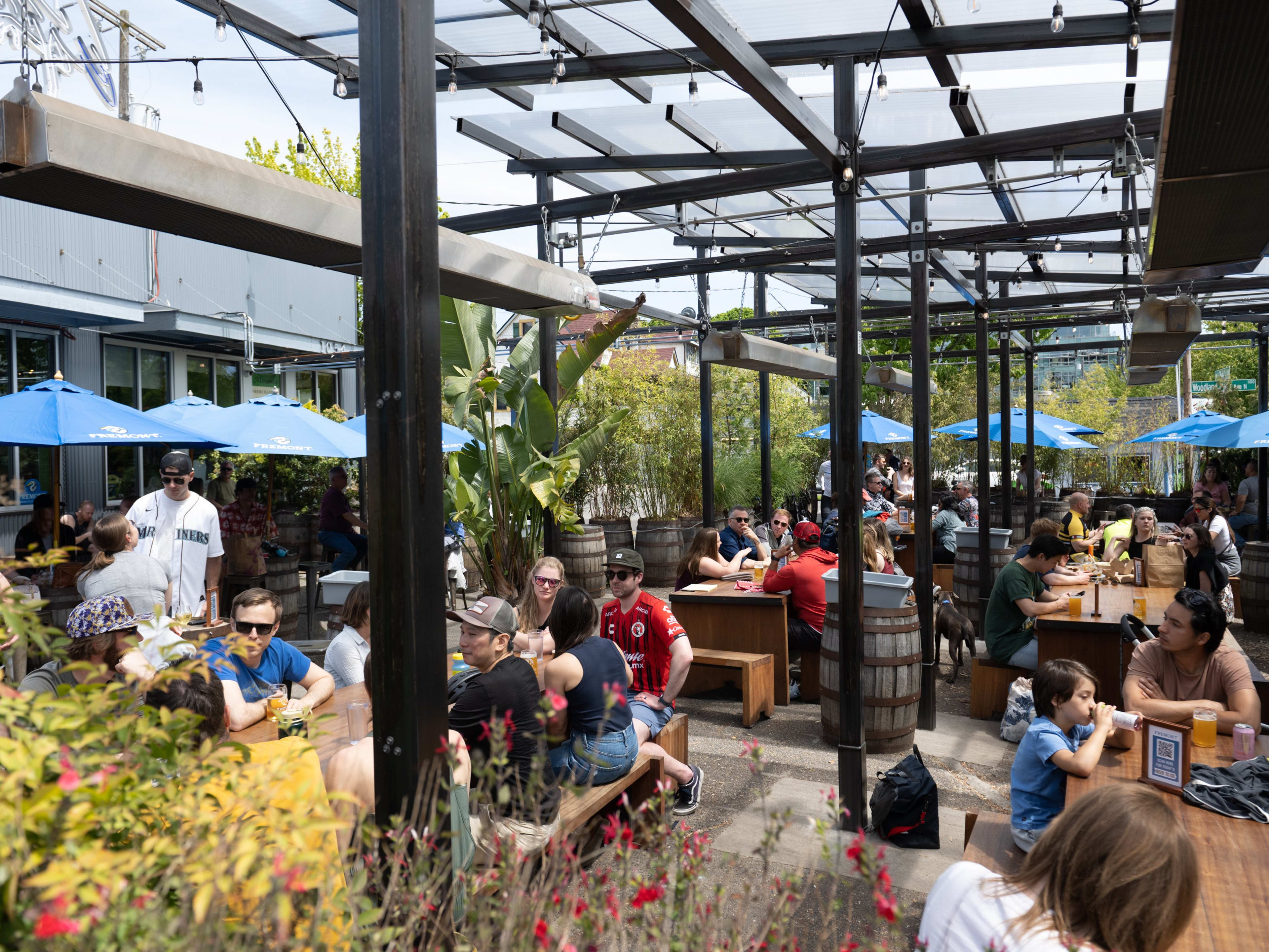 an outdoor beer garden with barrels, picnic tables, umbrellas, and people drinking beer.