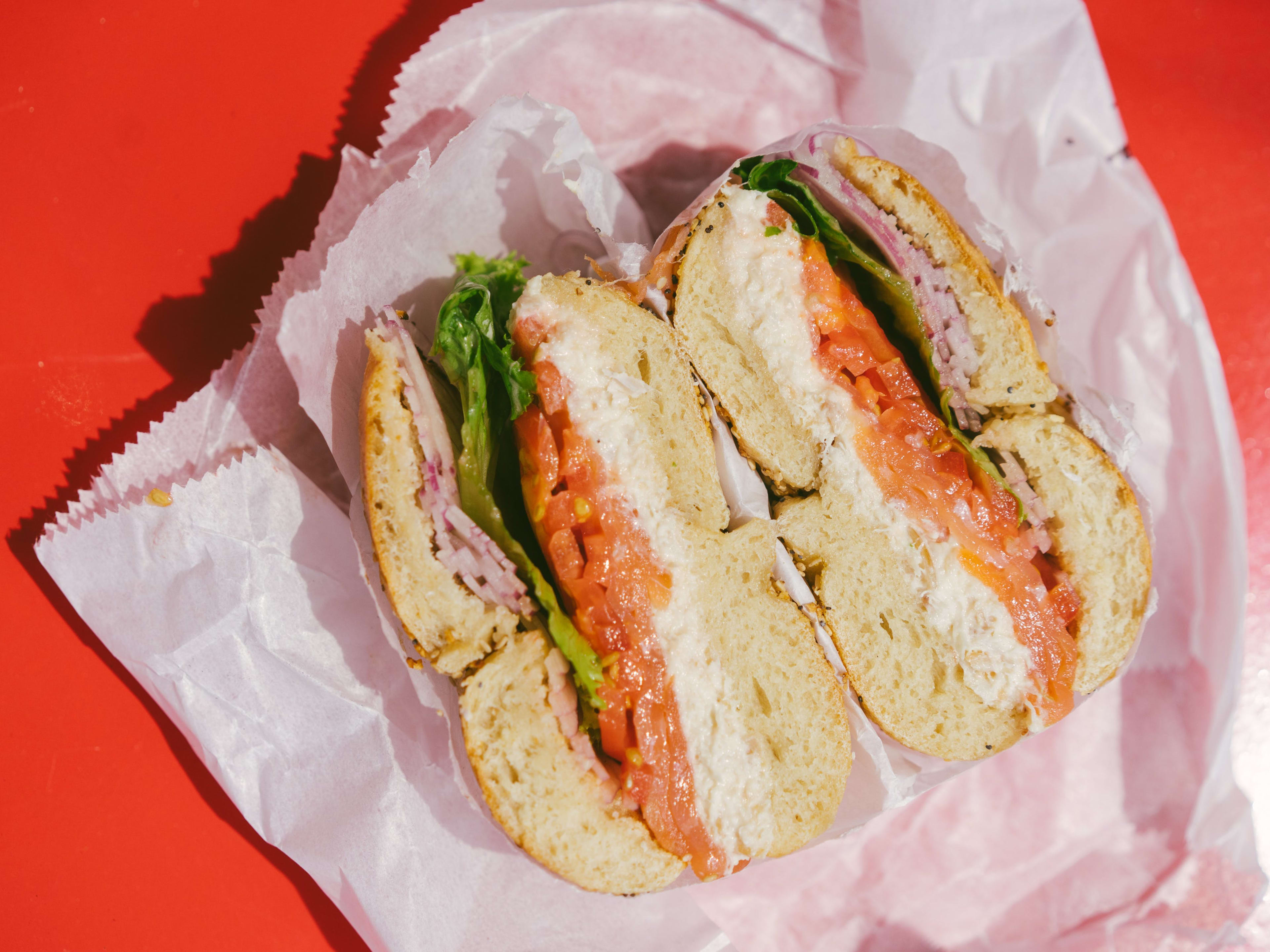 A bagel with cream cheese, lox, lettuce, tomato and red onion cut in half on a paper bag sitting on a red table.