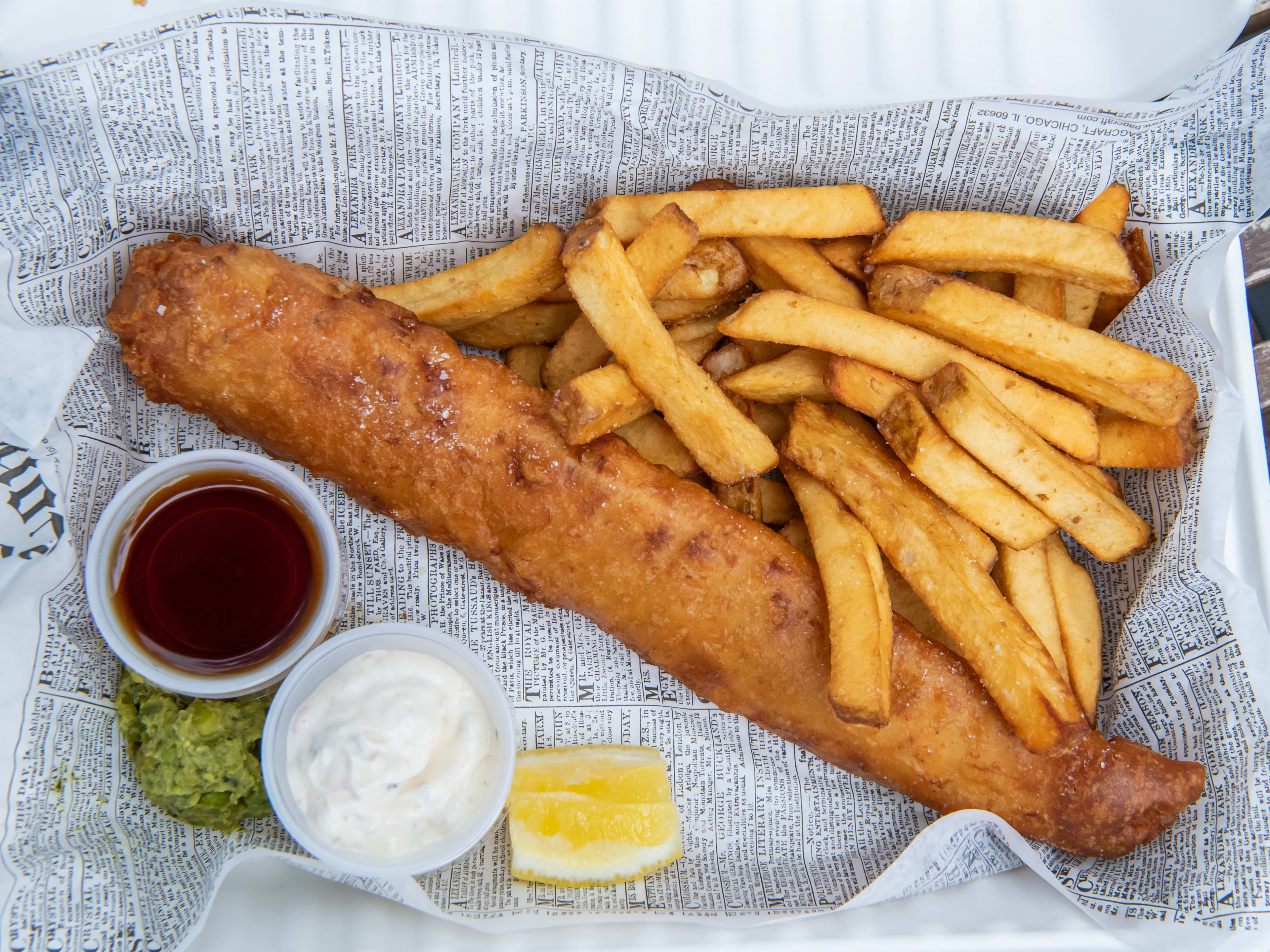 British-style fish and chips on a newspaper-lined tray with tartar sauce, malt vinegar, mushy peas, and a lemon wedge.