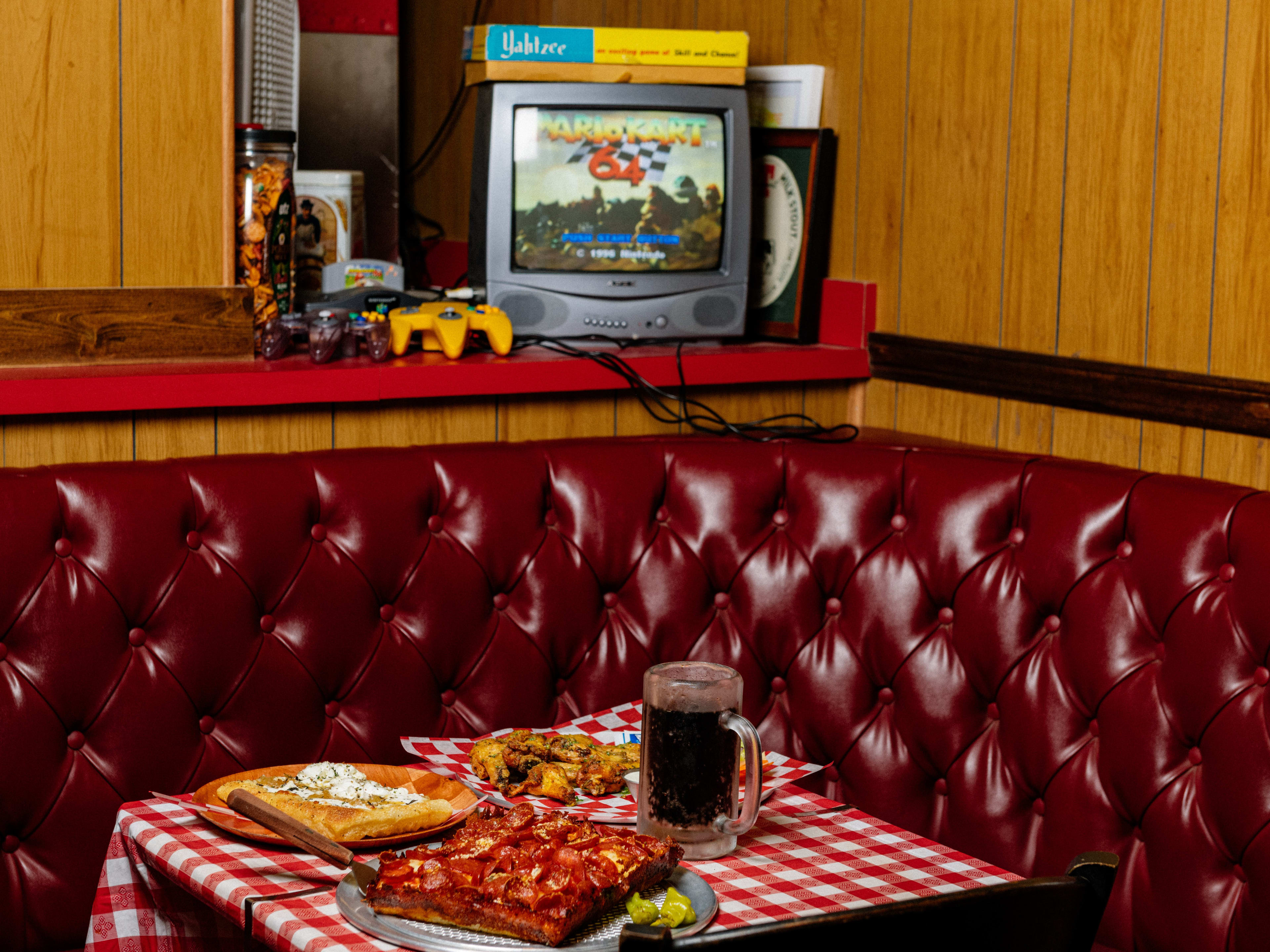 A pepperoni Detroit style pizza, a grandma pizza, wings, and a soda in a glass mug on a checkered table cloth at a red vinyl booth. A ledge behind the booth has a TV with N64 Mario Kart and other retro games.