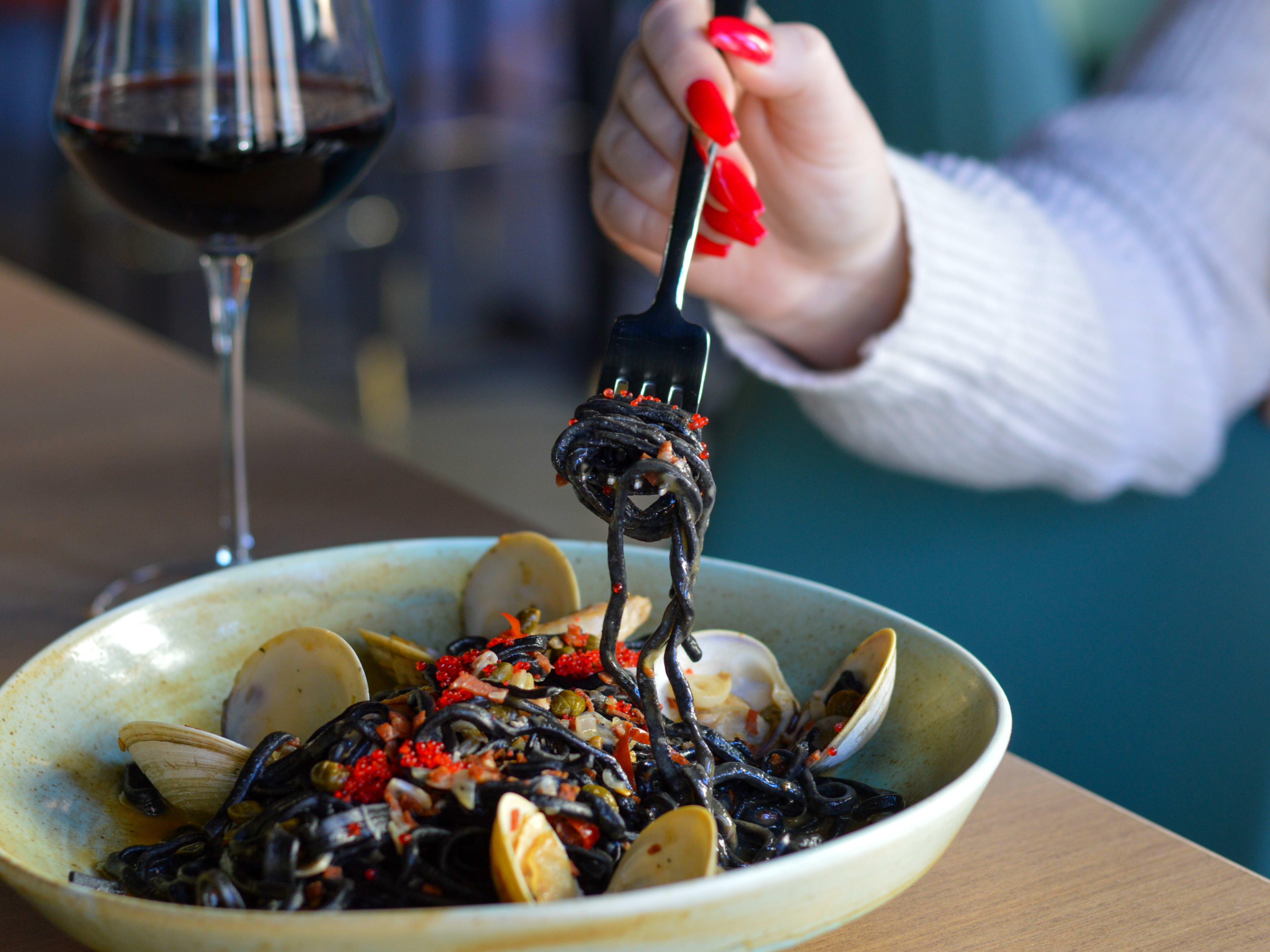 A person twirls squid ink pasta on a fork. The rest of the pasta and clams are served in a ceramic bowl. A glass of red wine is set to the side.