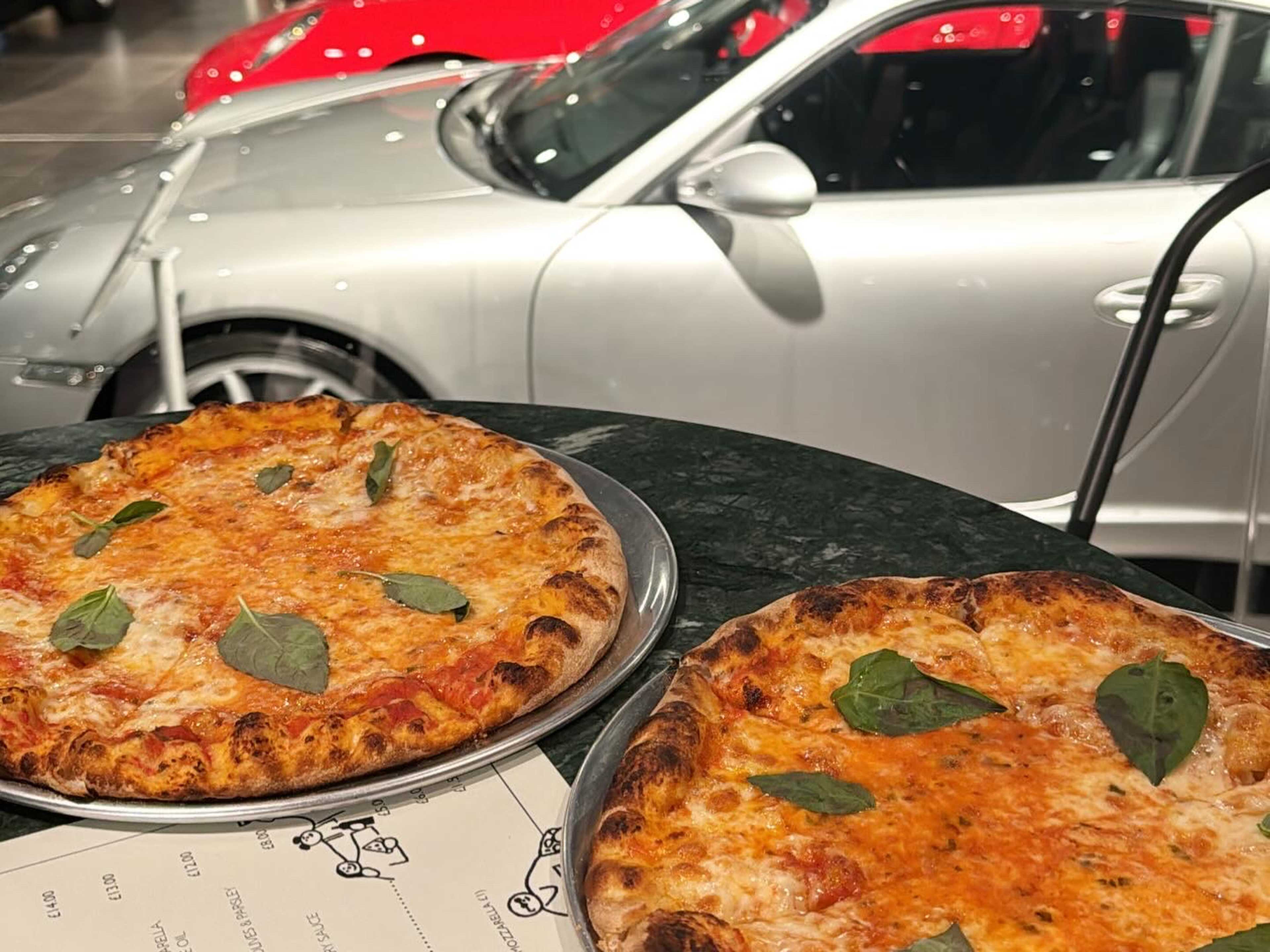 Two pizzas on a table, with vintage cars in the background.