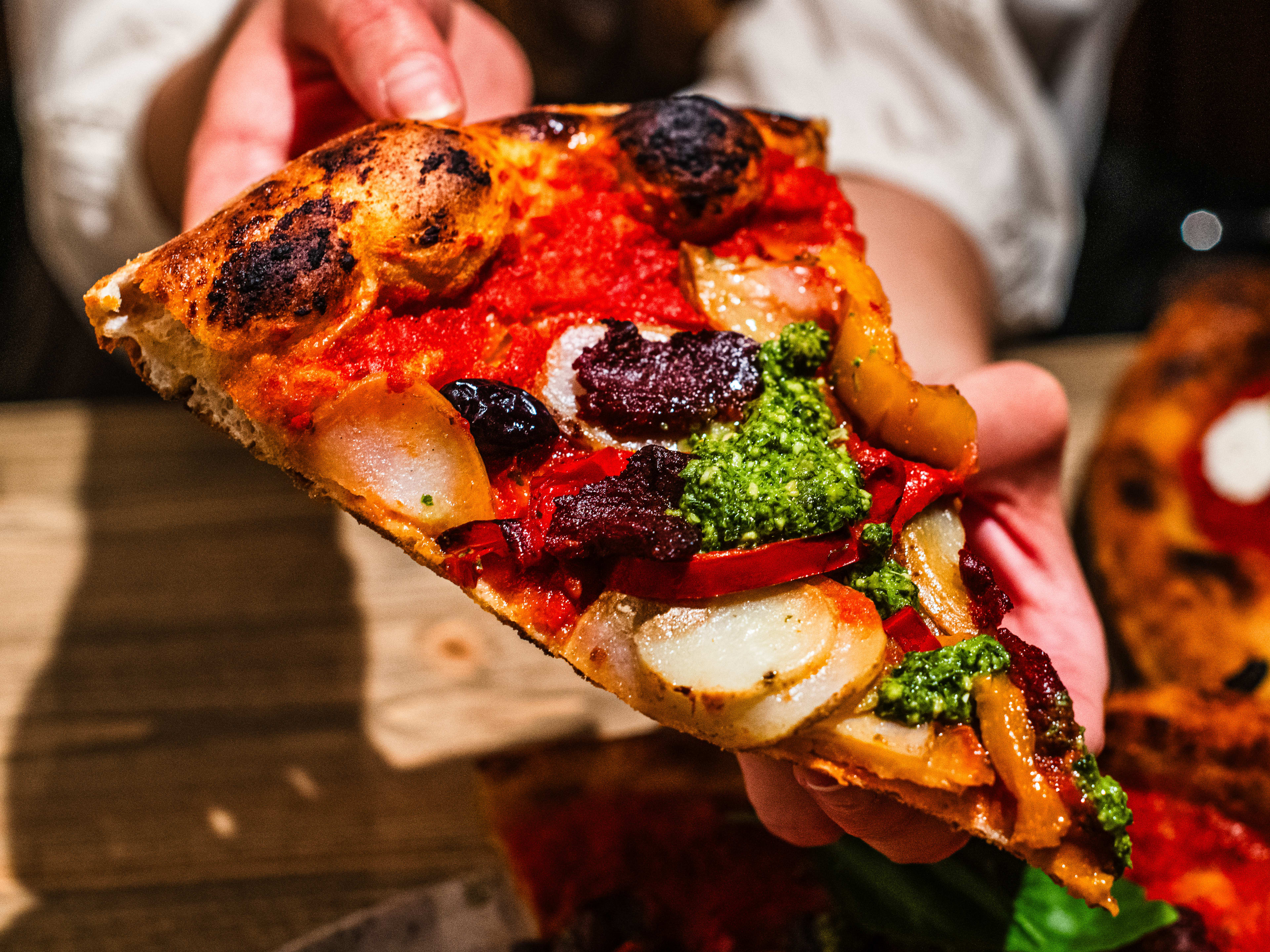 A person holding up a slice of the Vegan N' Fire pizza from Alley Cats Pizza.