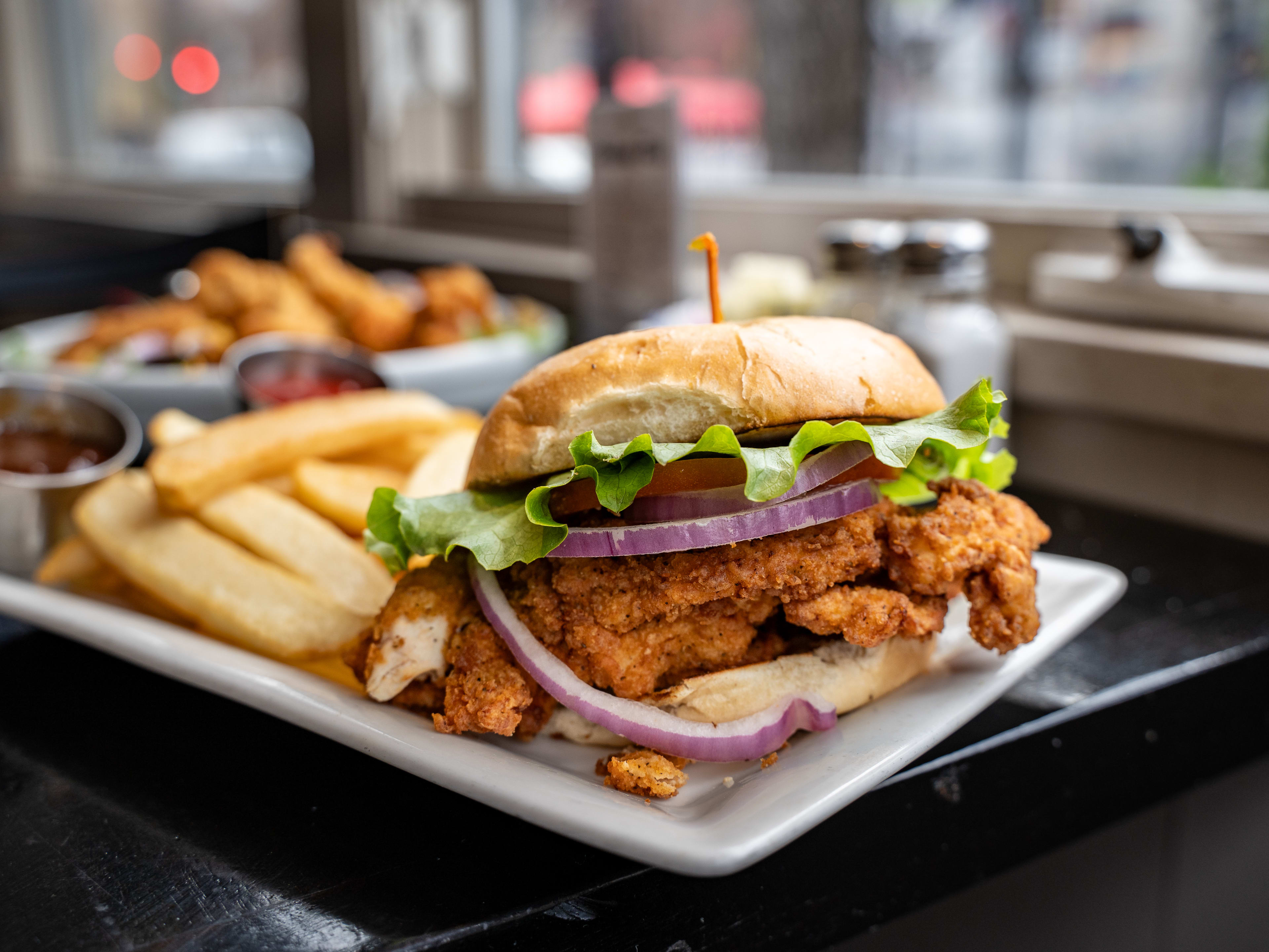 Fried chicken sandwich topped with red onion, lettuce, and tomato served next to steak fries and sauce.