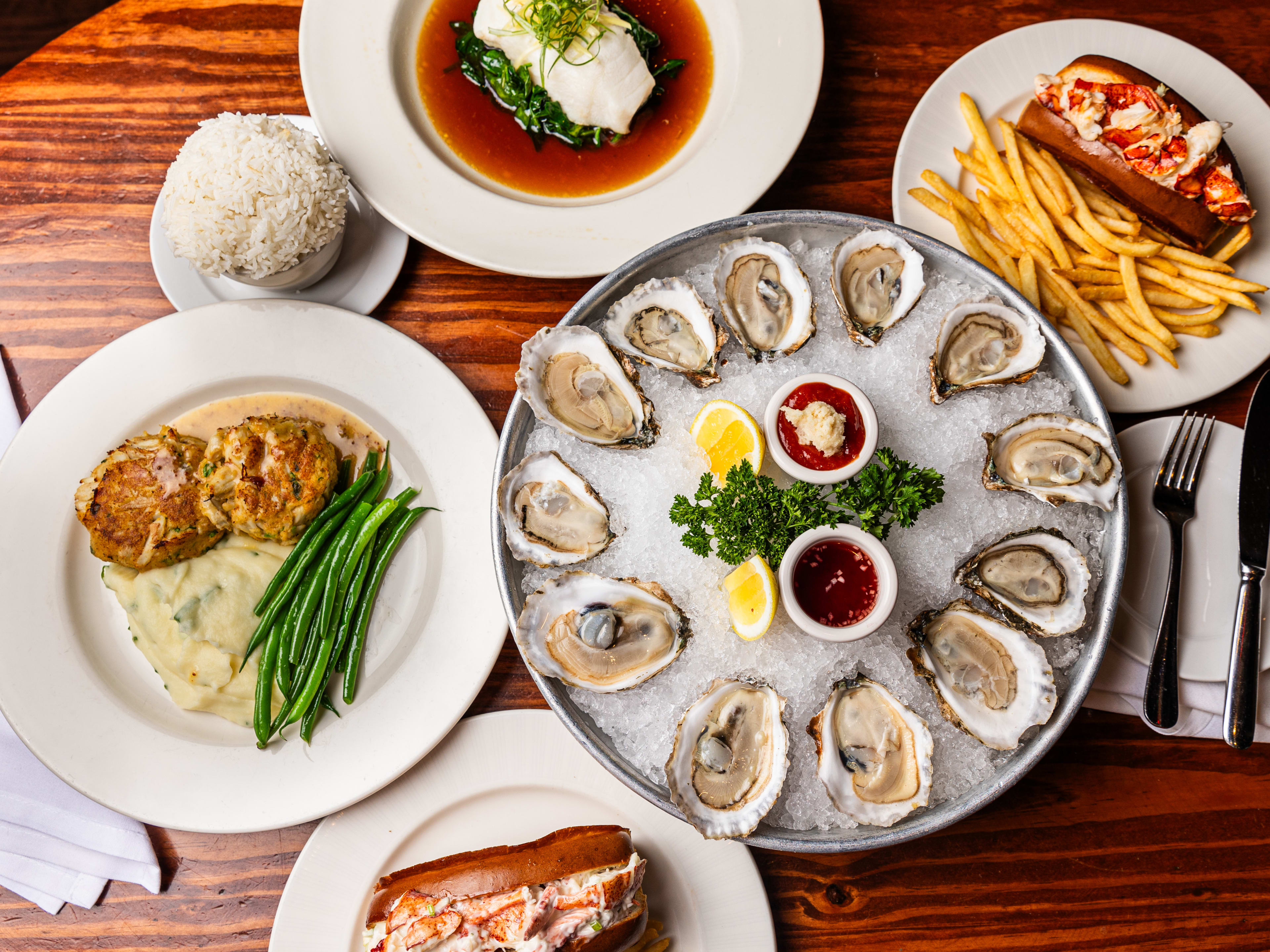 A food spread with oysters, a lobster roll and fries, and white fish.