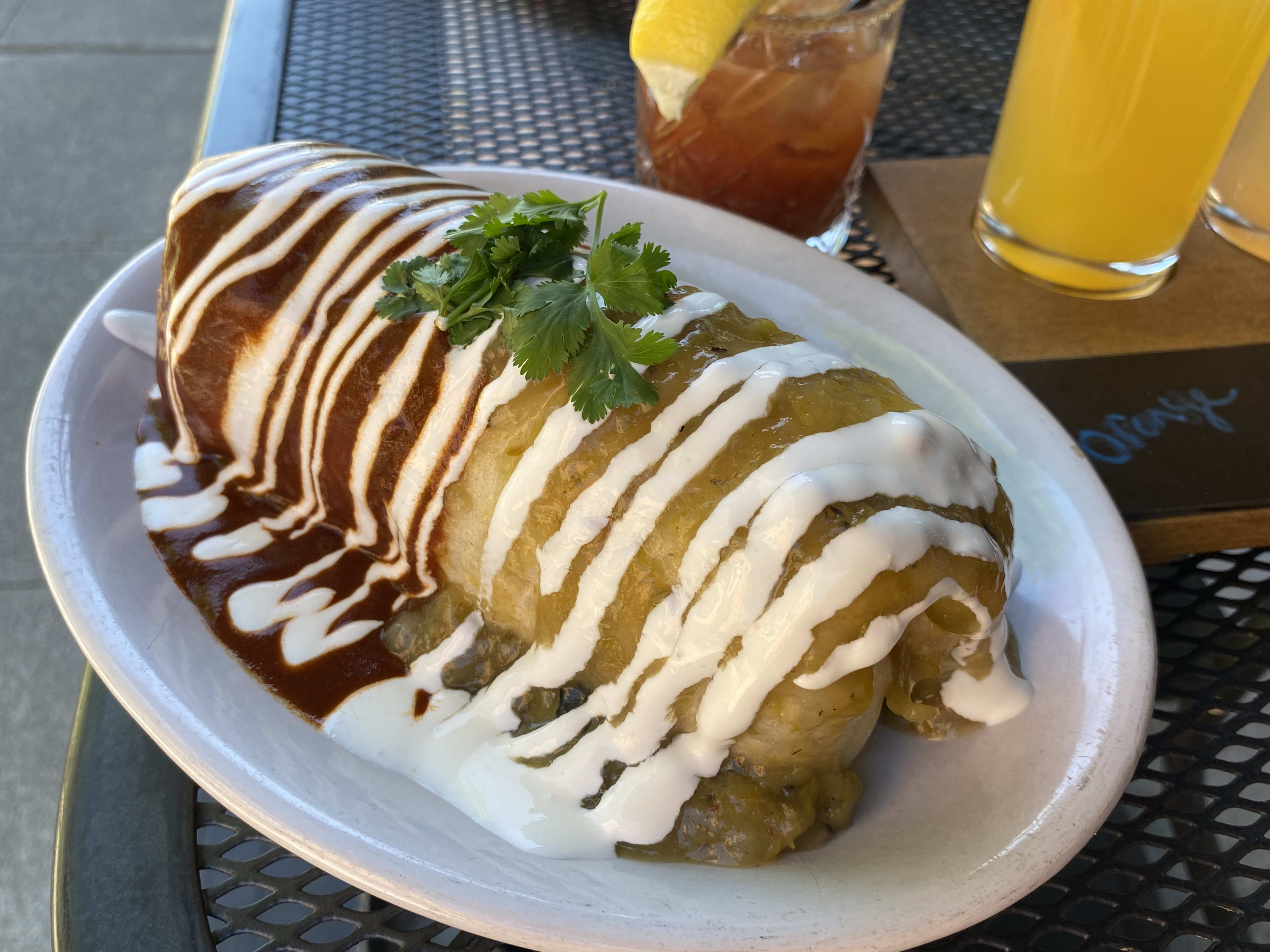 A large breakfast burrito drenched in red and green sauce next to a Bloody Mary and flight of mimosas