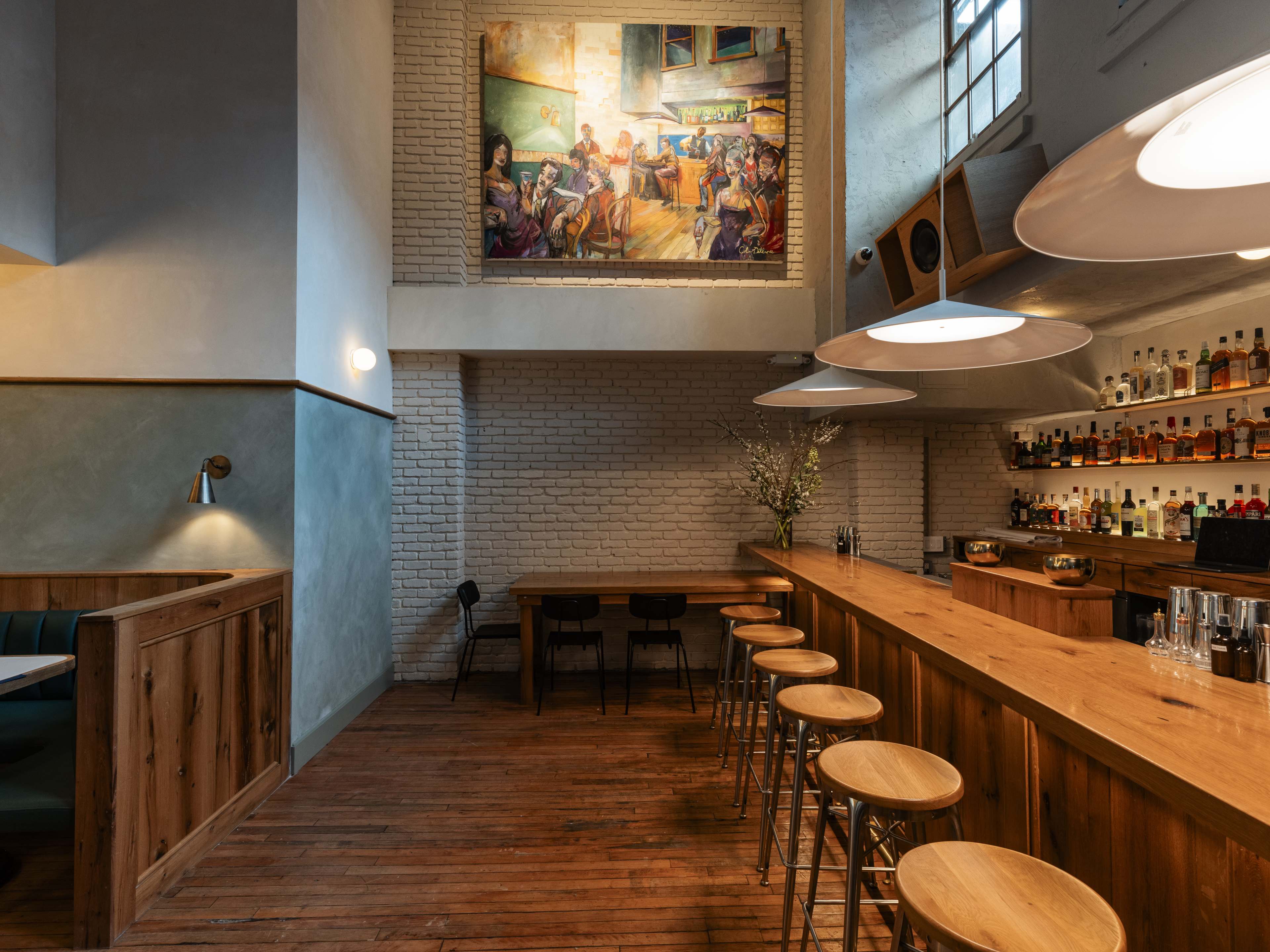 A cocktail bar with a wooden bar, high ceilings, and a painting of a cocktail bar on the wall.