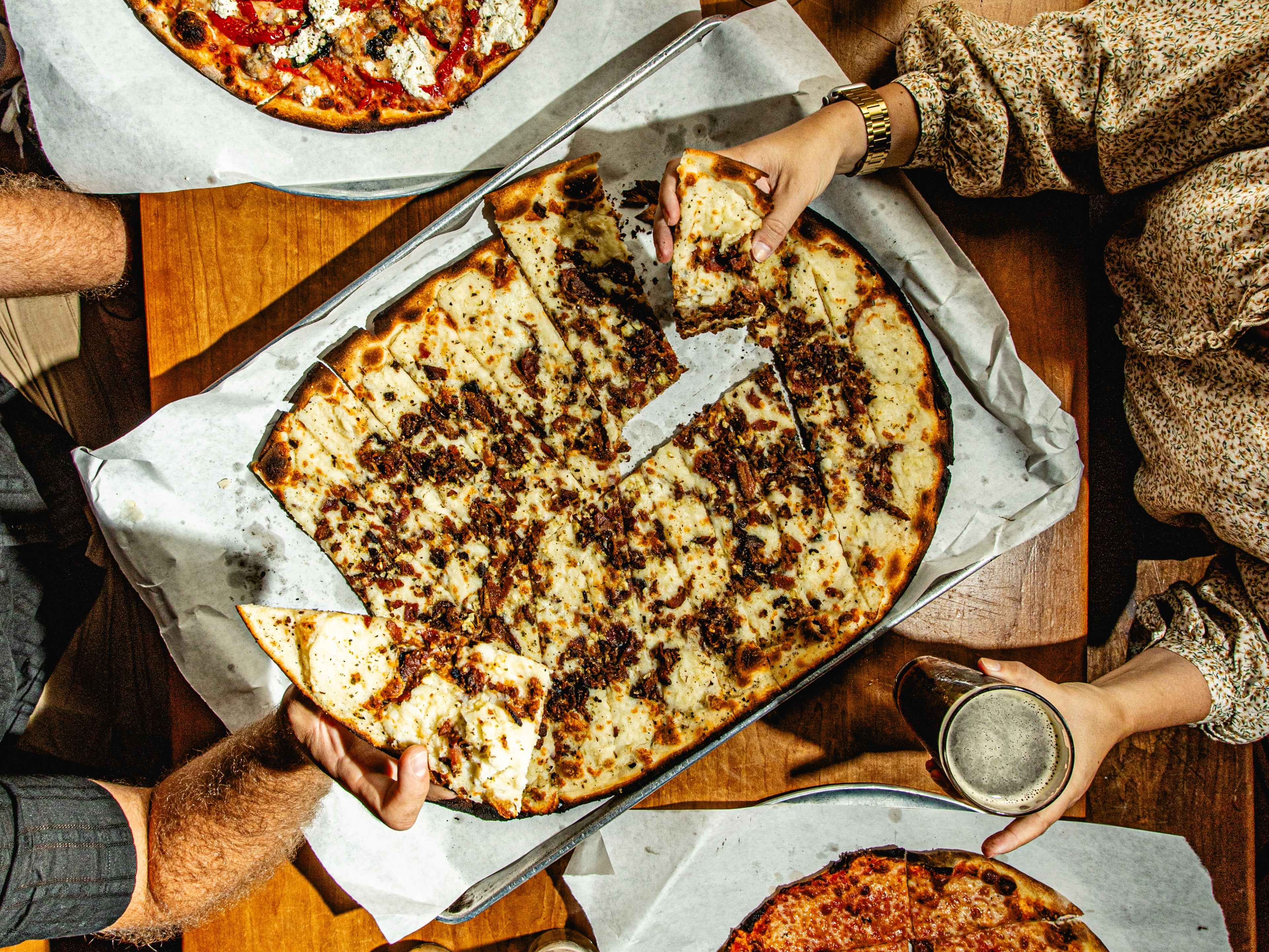 Two people reaching over a table to get a slice of pizza