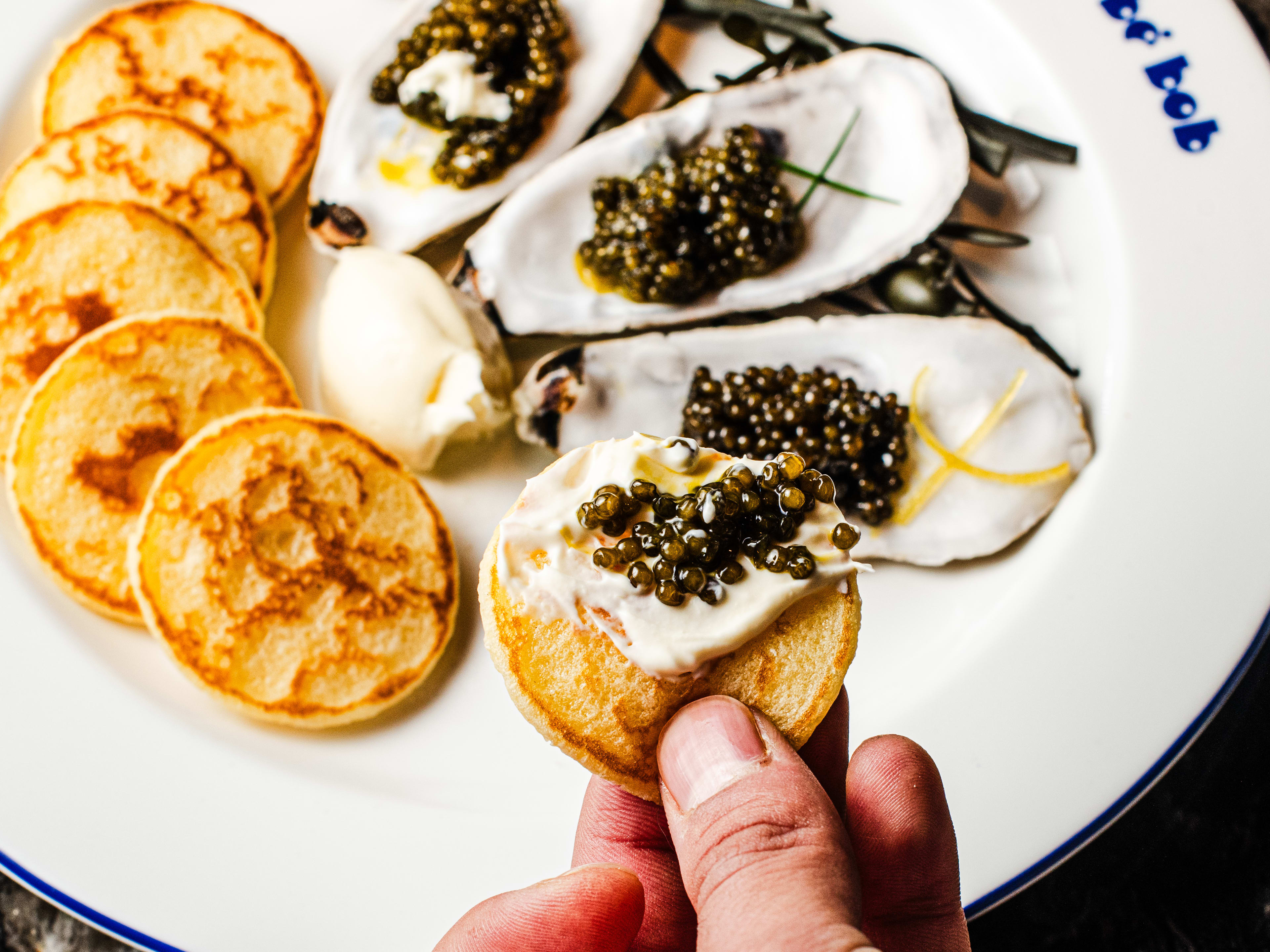 Three oyster shells filled with caviar, butter, and blinis on a plate. One blini is held up with butter caviar spread on top.