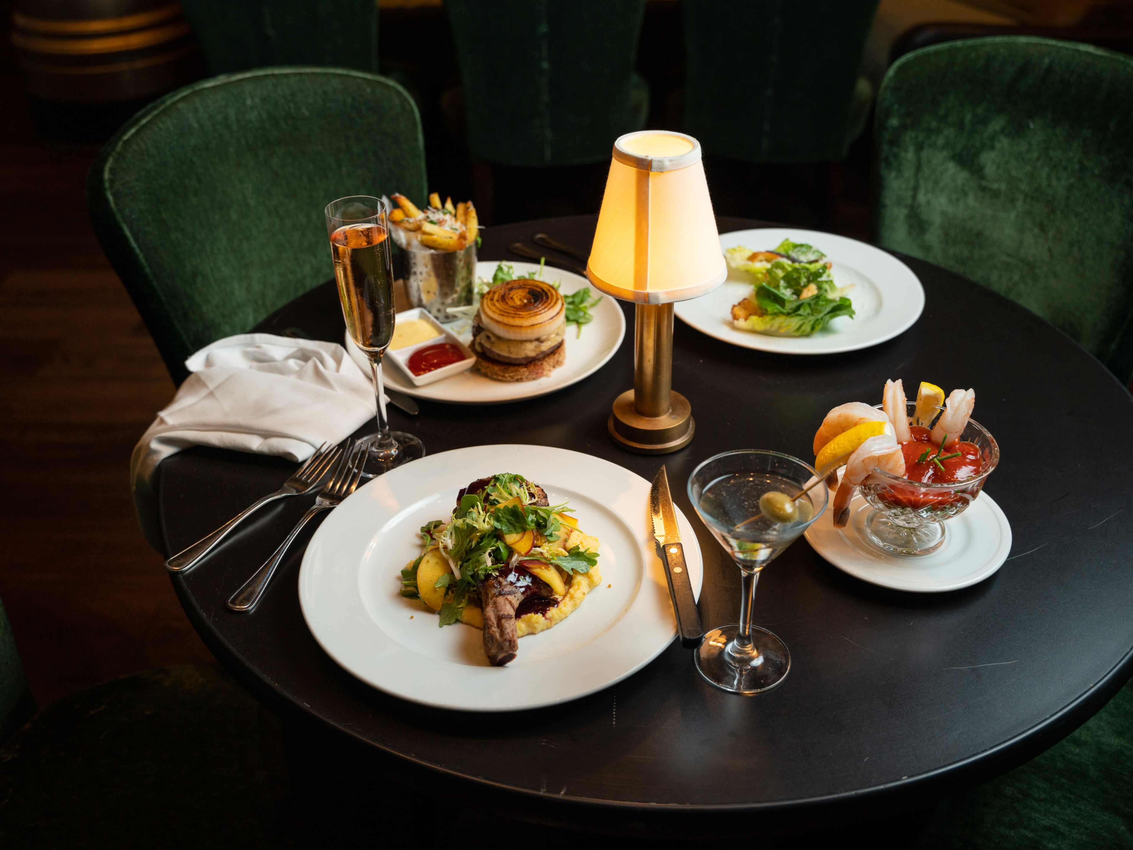 spread of burger, salad, shrimp, cocktail, and martini on table with tiny lamp in the middle and velvet chairs surrounding