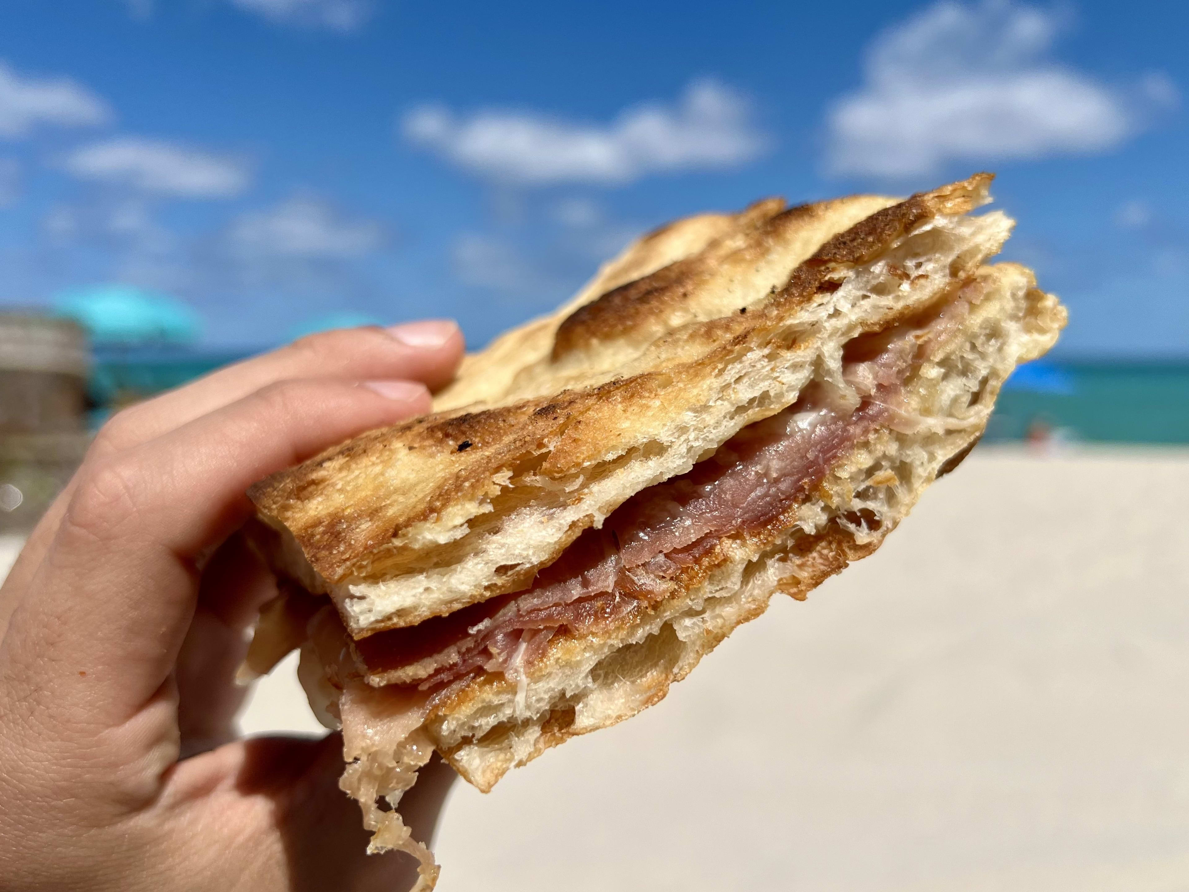 A sandwich held up on the beach, with the ocean in the background.