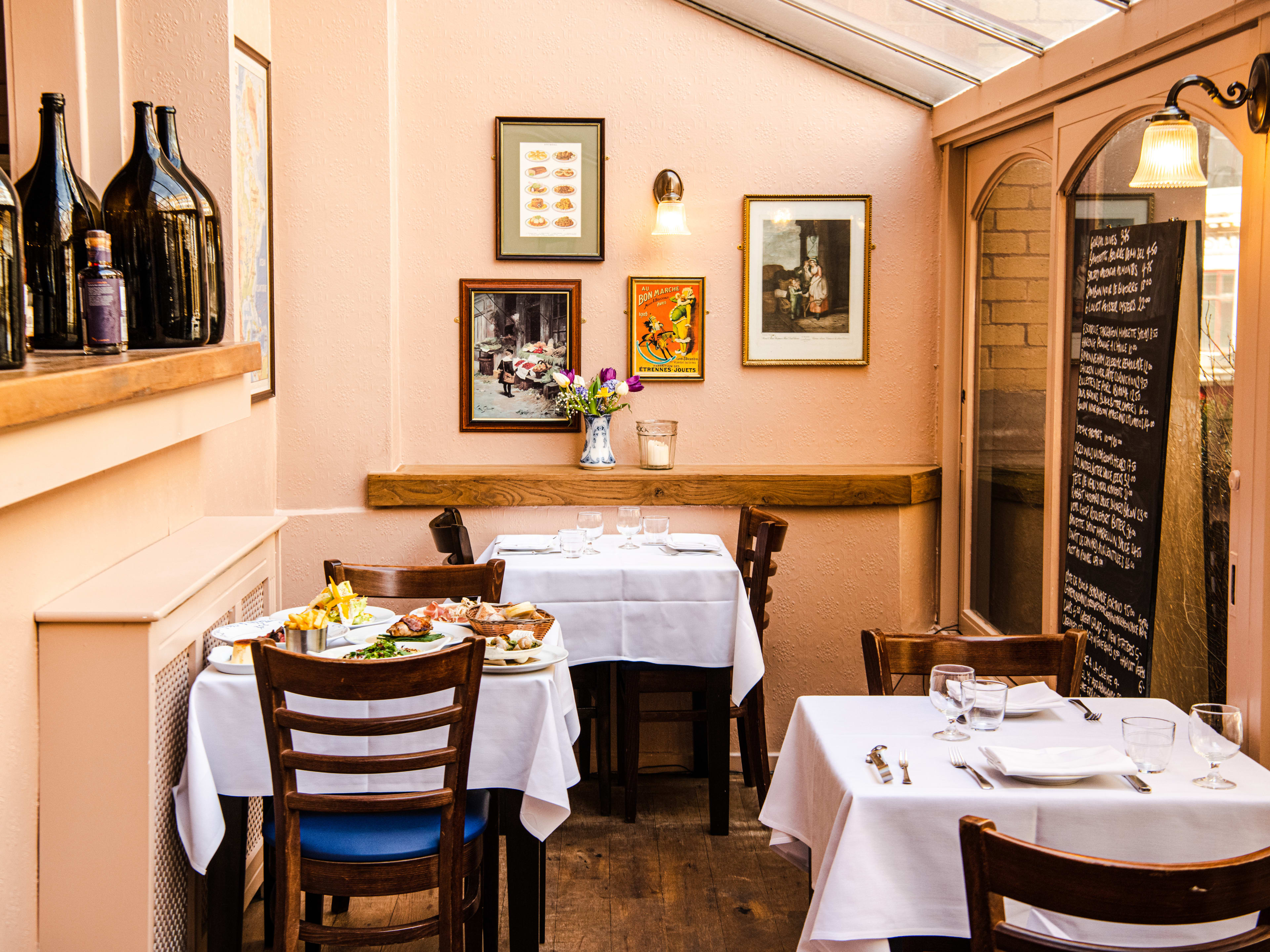 A corner of Bouchon Racine with three two-person tables, a chalkboard menu on the walk, and gold-framed pictures hanging on the wall.