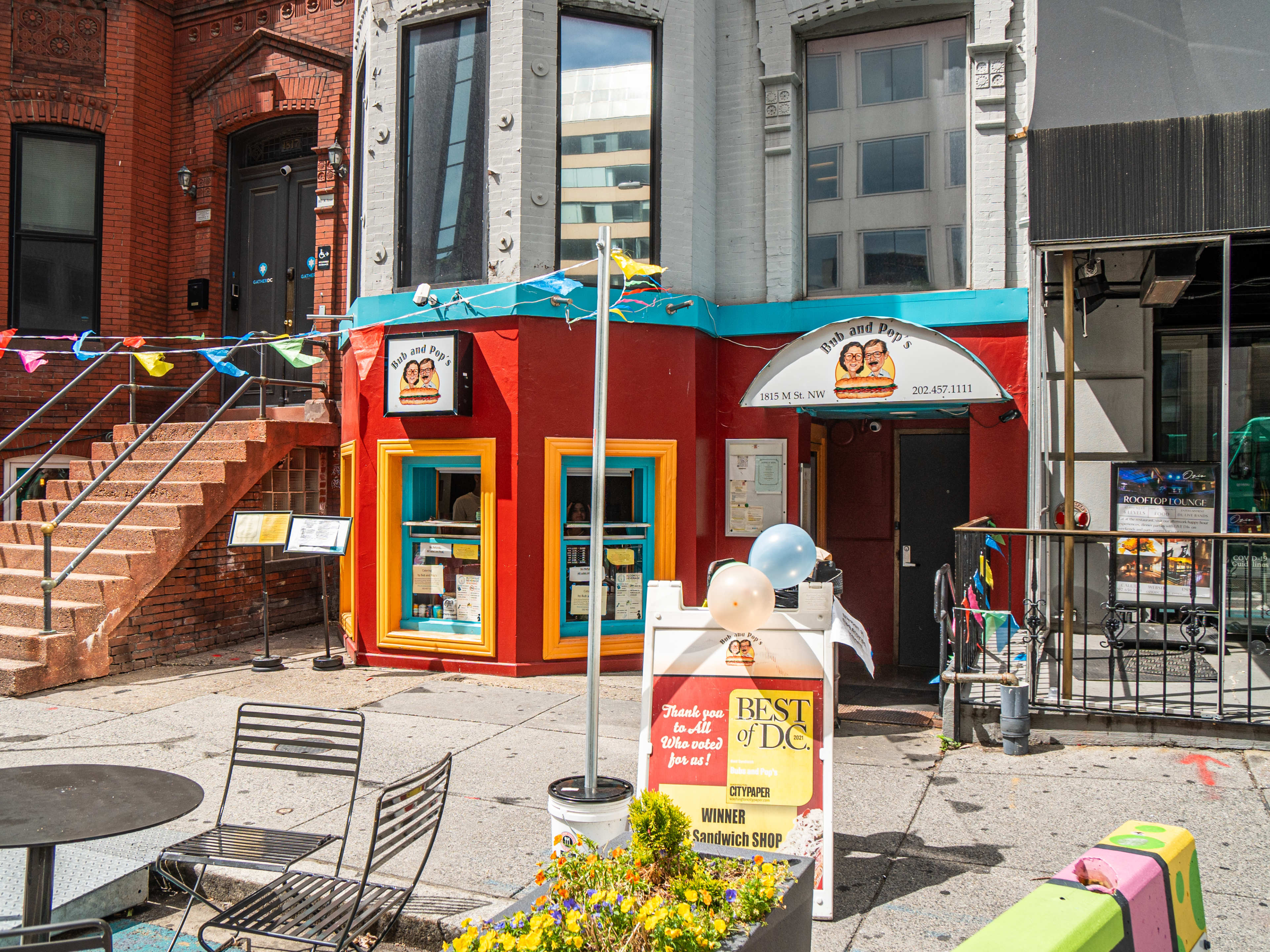 colorful exterior of deli with flag banners hanging across sidewalk and outdoor seating setup