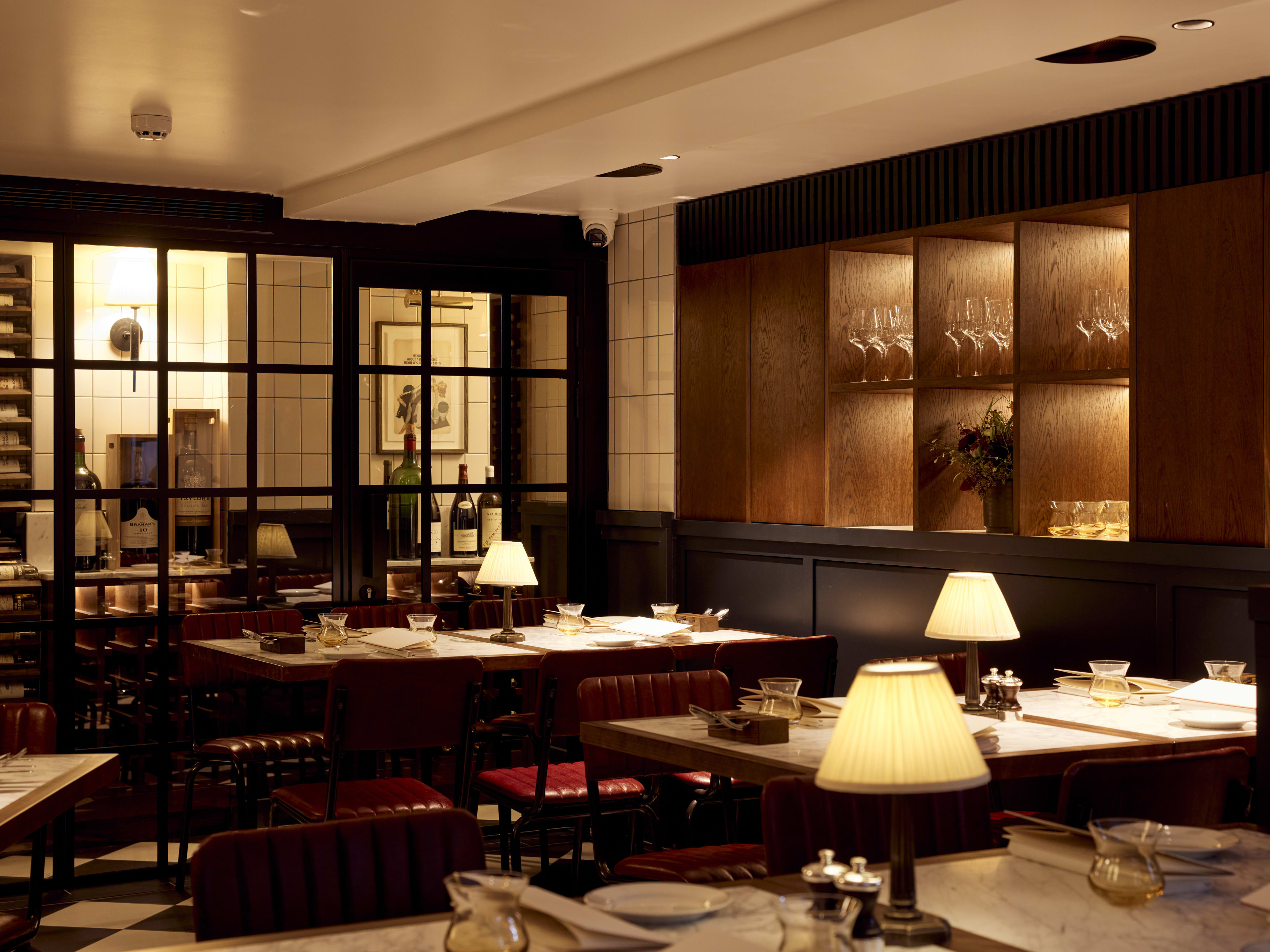 Restaurant interior with moody lighting, mini lamps on each marble-look table, and oxblood chairs.