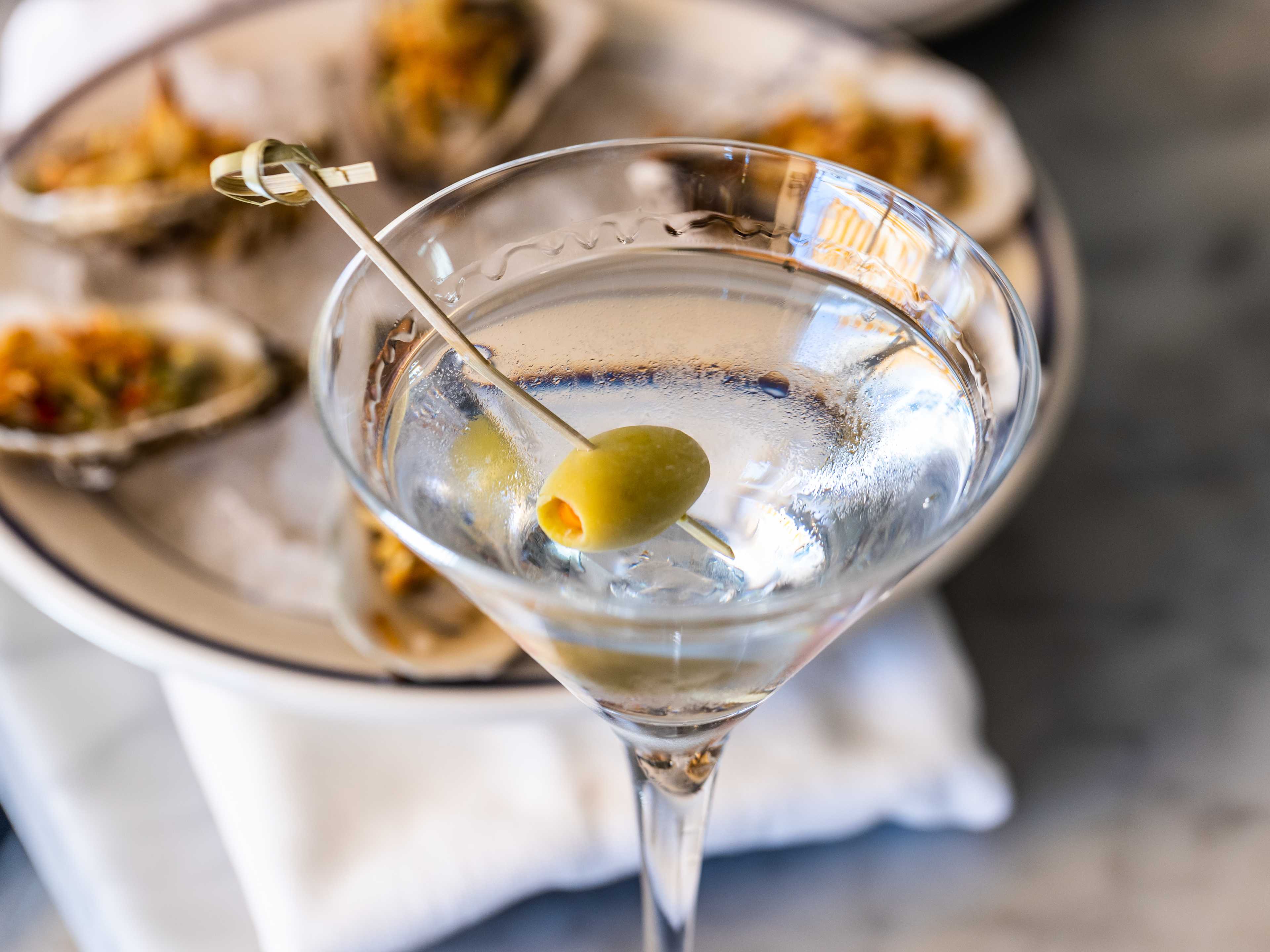 A gin martini with an olive. Oyster on a plate in the background.