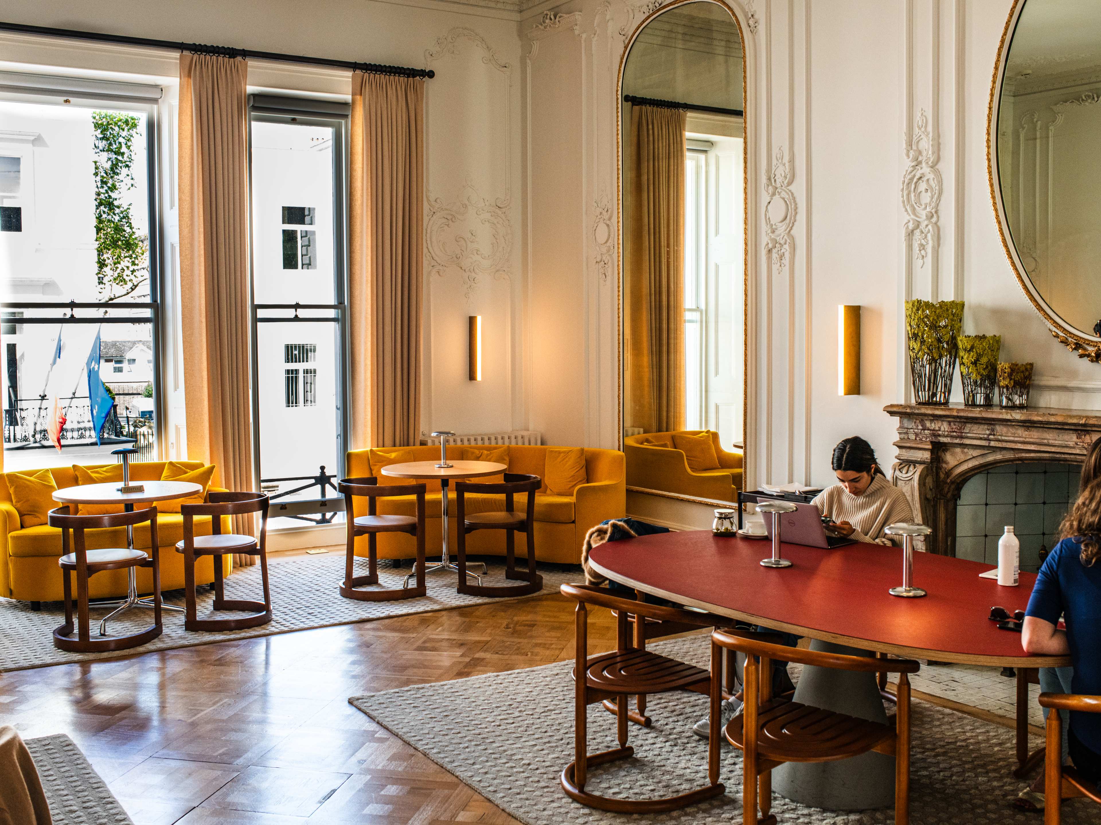 A cafe with stuccoed walls, a communal table with people working, and yellow booths in front of a window.