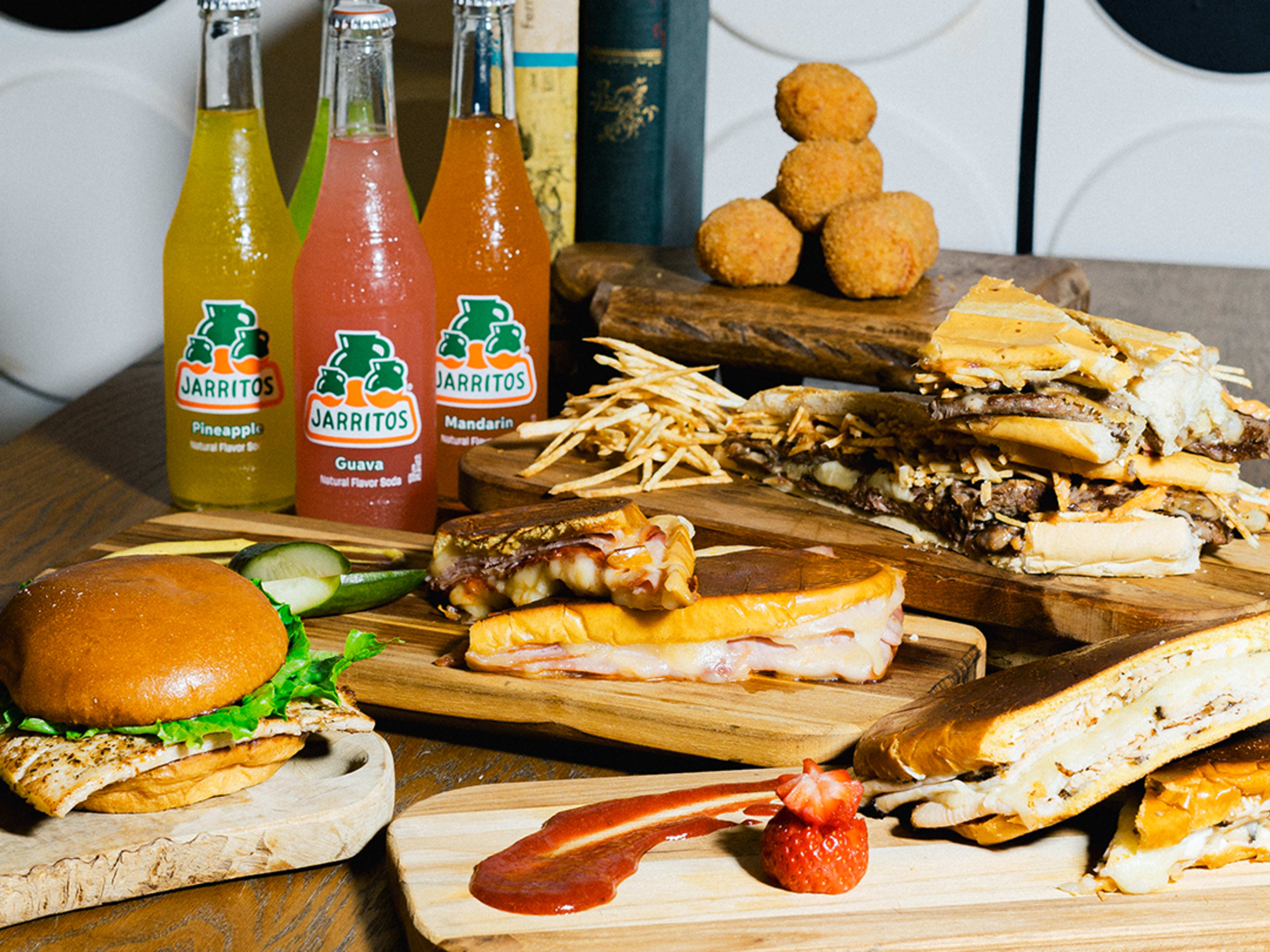 a display of food including four bottles of Jarritos soda, three toasted sandwiches on cuban bread, a tower of round croquetas, and a mahi sandwich