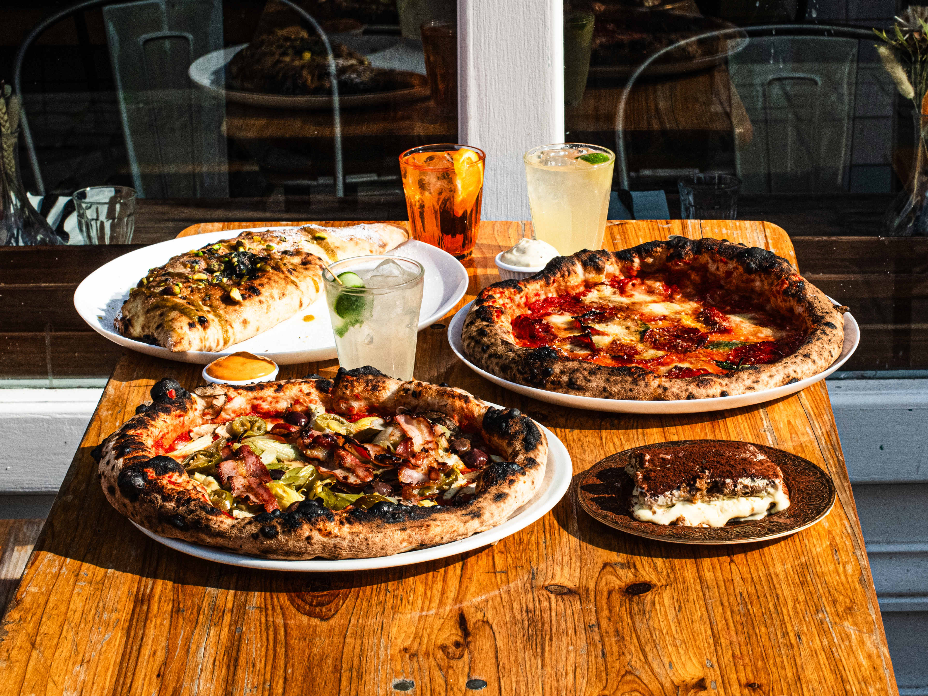 Two pizzas, a tiramisu, drinks, and a calzone on a wooden outside table in the sunshine.