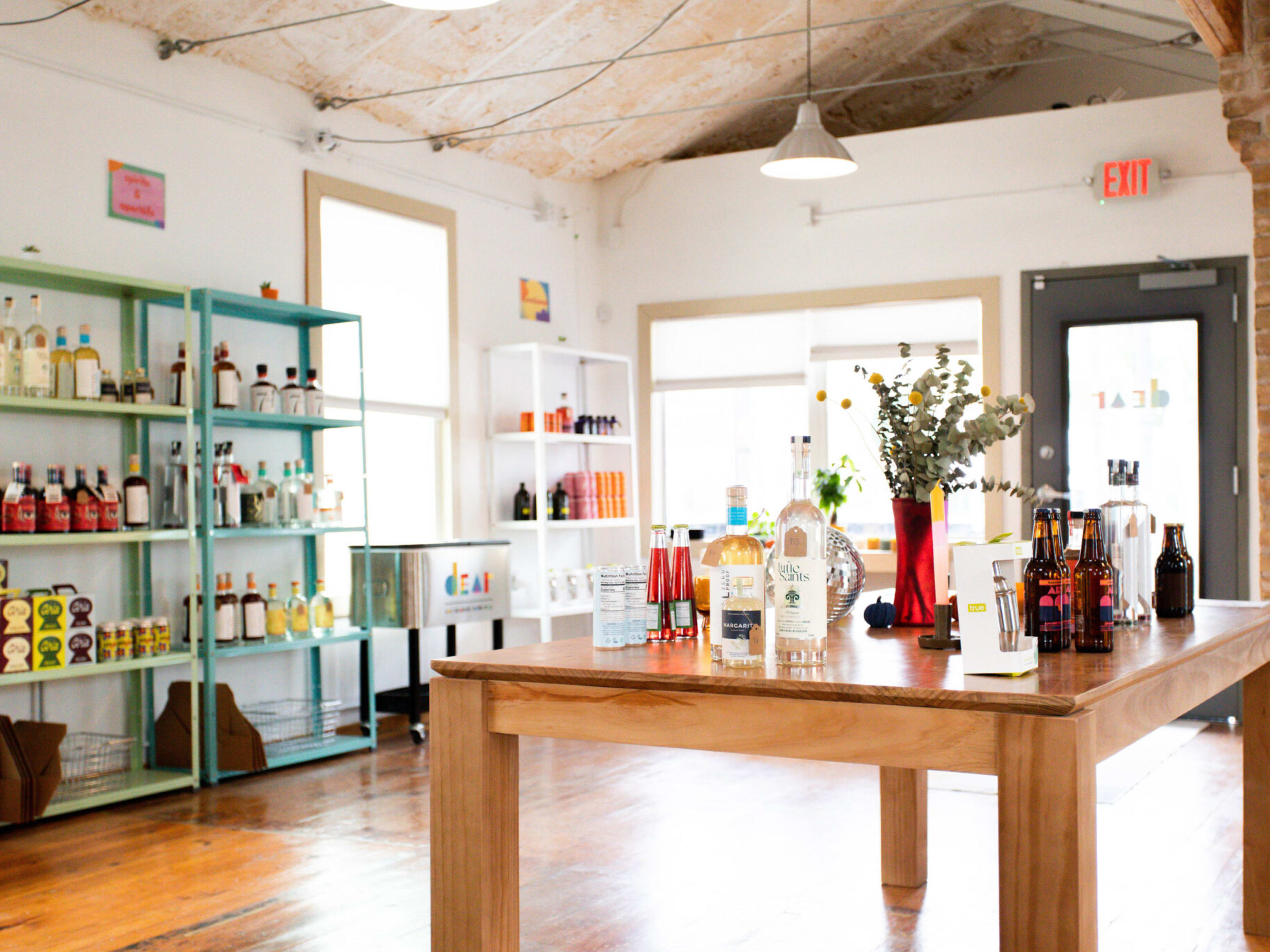 The interior of Dear Dry Drinkery with non alcoholic bottles on display on shelves and a wooden table in the center. There are also other small items for purchase.