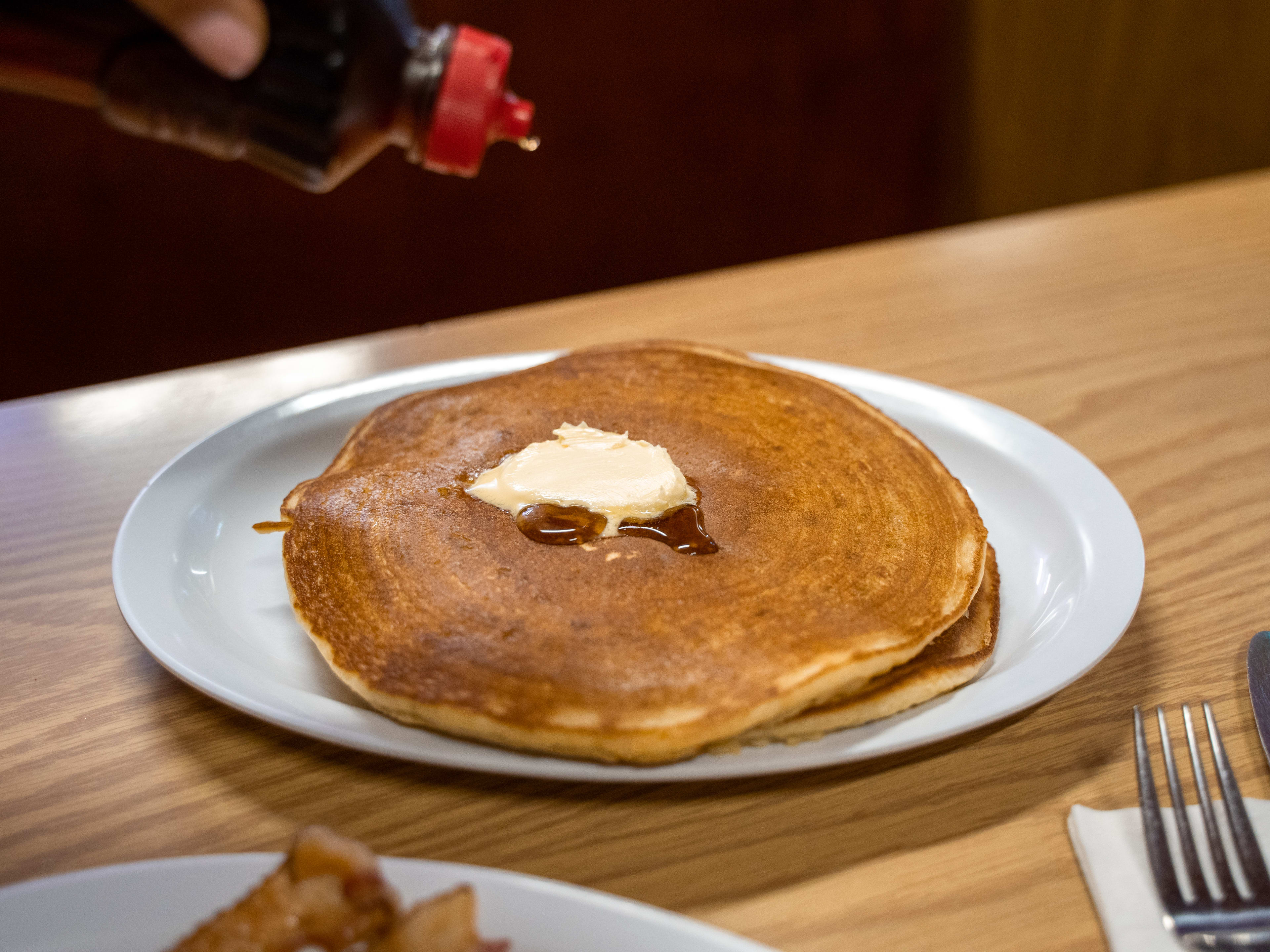 Two large flat pancakes on a diner plate with a pat of butter and syrup being drizzled over them.