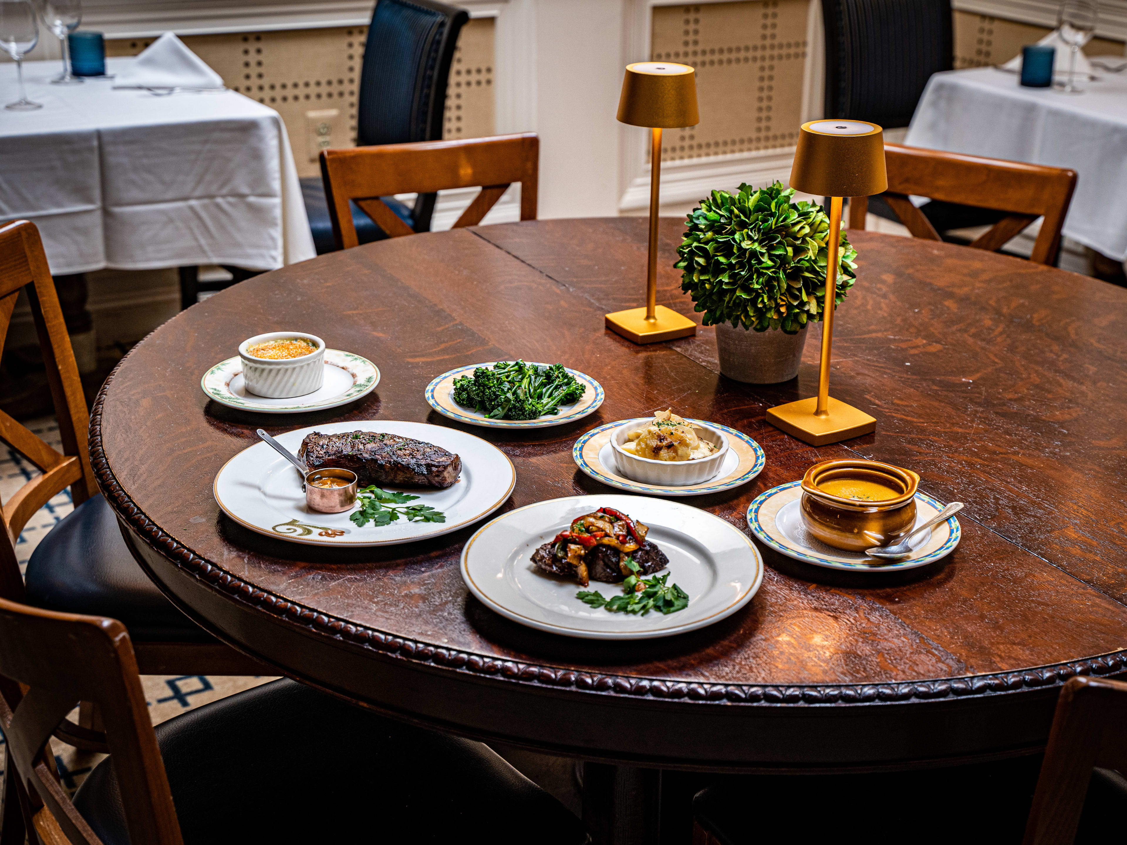 A spread of steaks and sides at an old school steakhouse with white tablecloths and lamps on tables.