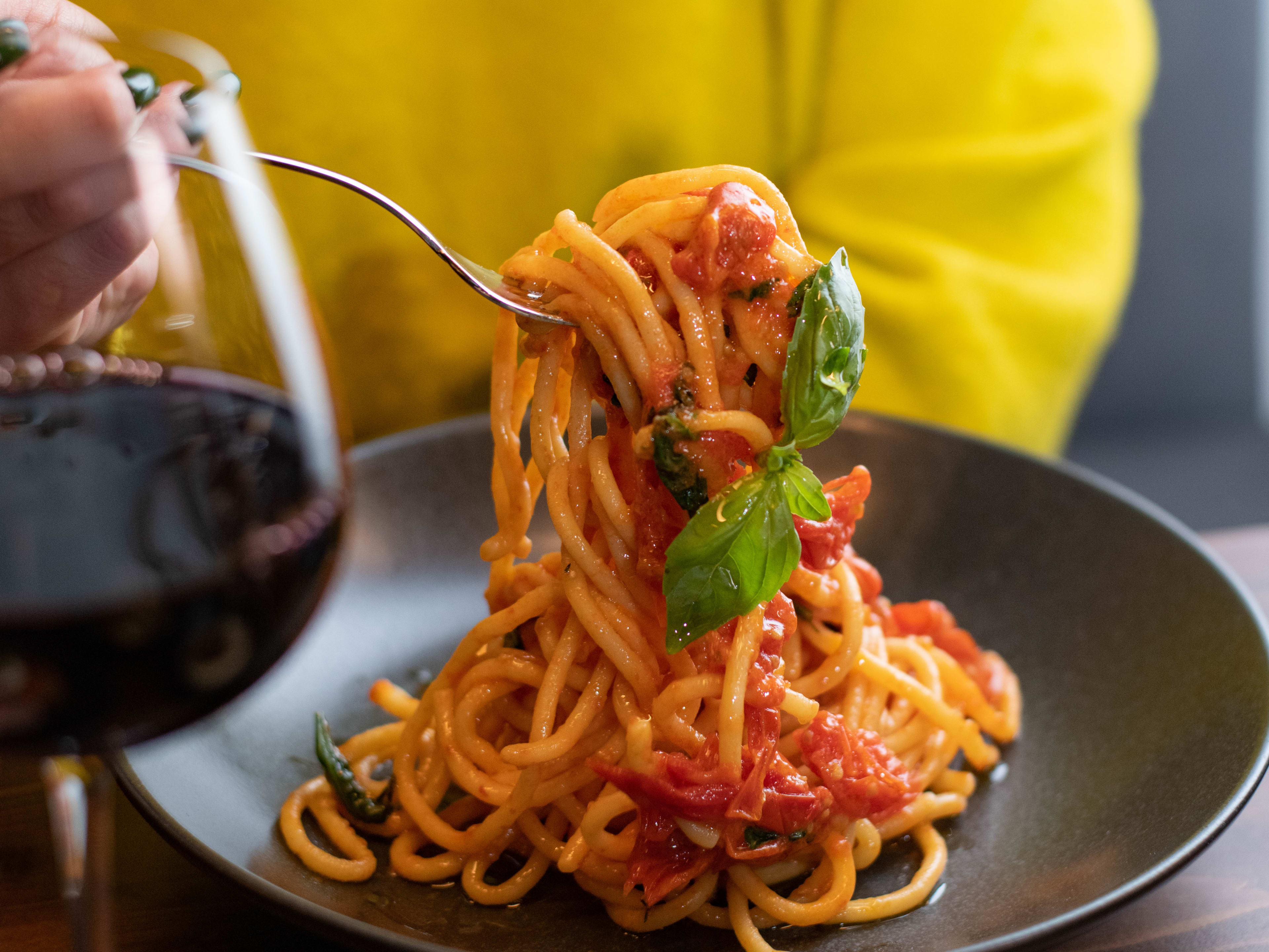 A person in a yellow sweater picks up spaghetti with a fork off of a grey plate. A glass of red wine is also on the table.