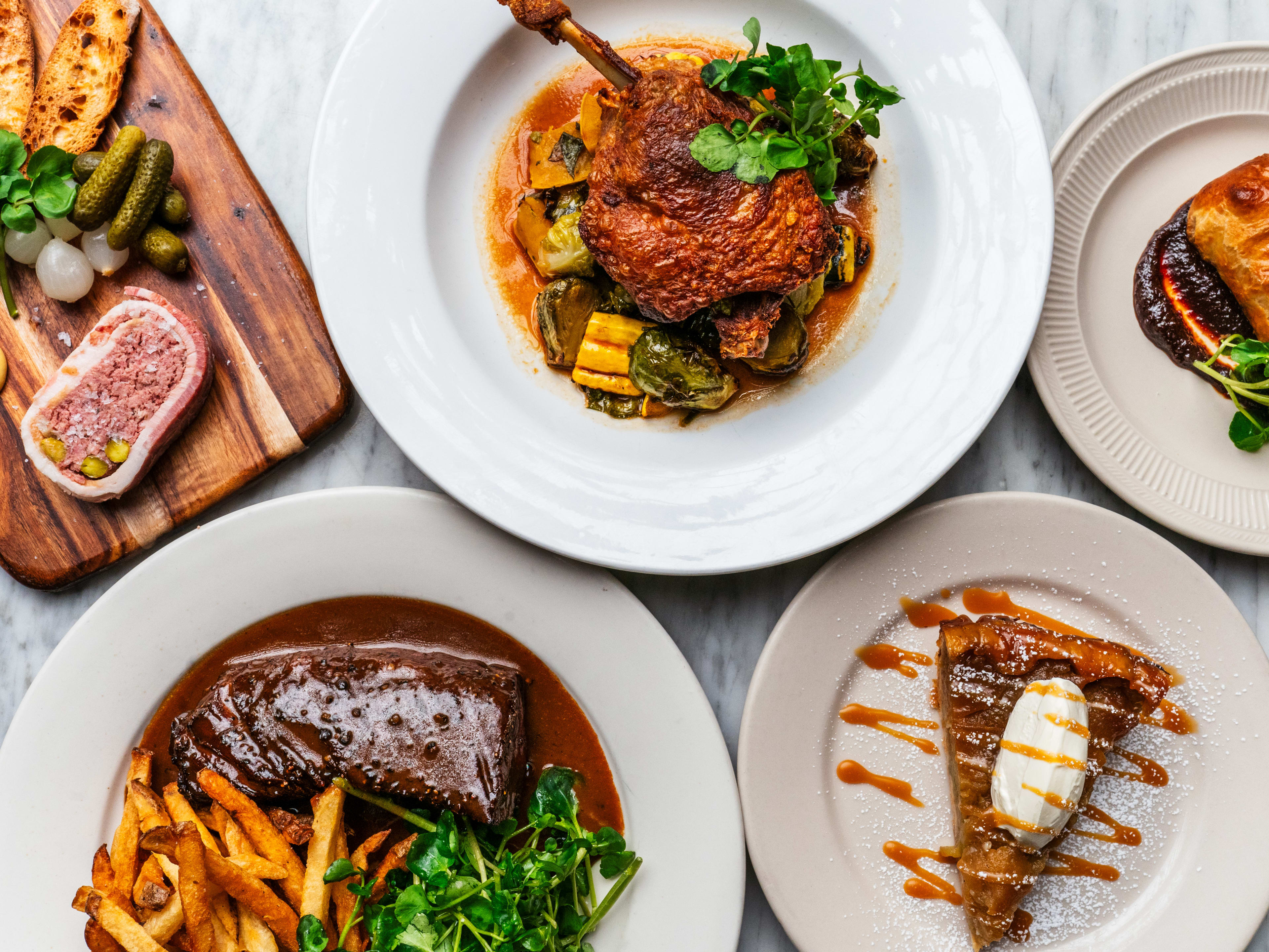 A spread of food on a marble table, including roast chicken, pie, and steak frites.