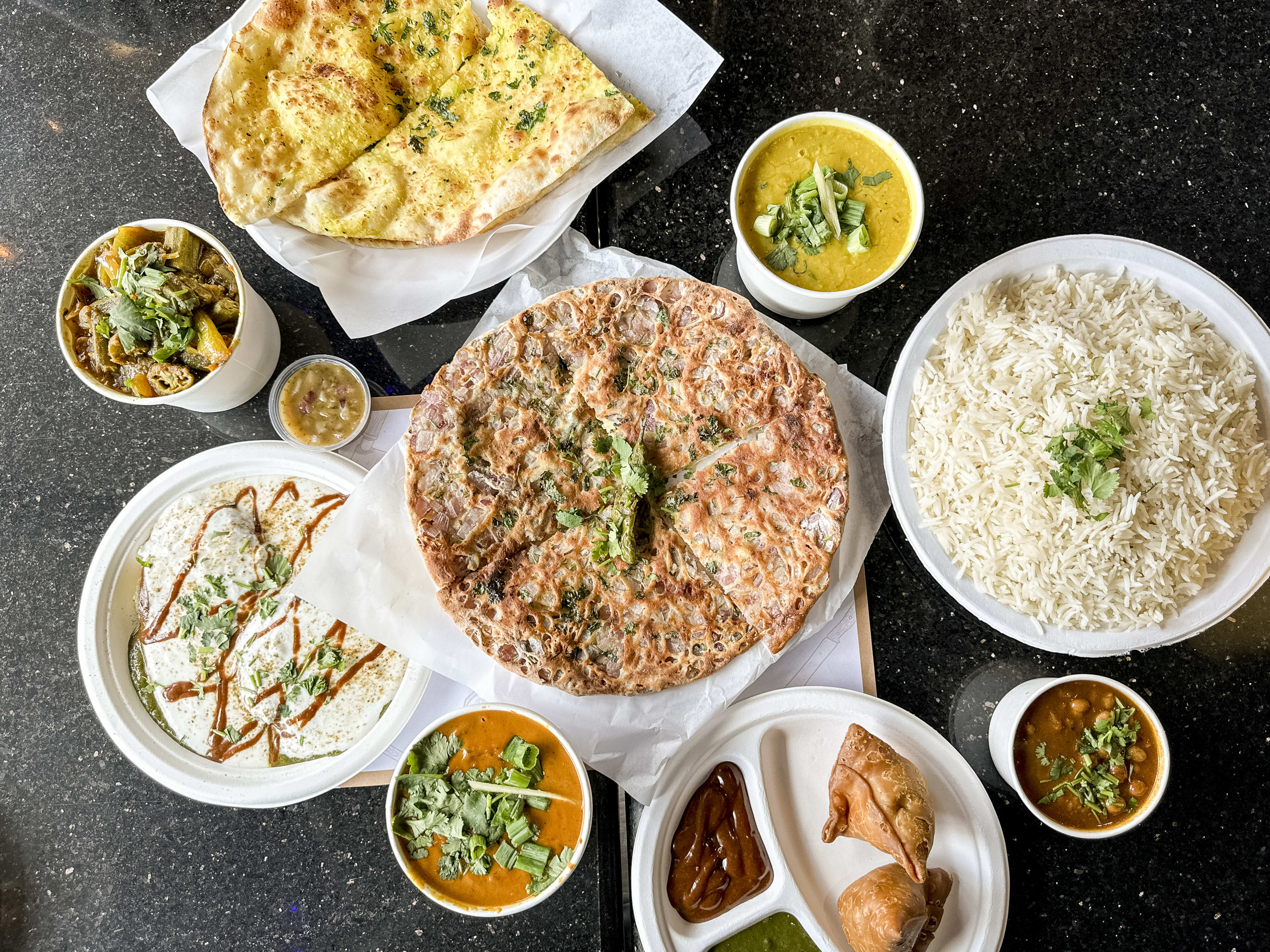 a tabletop spread of Indian dishes, like basmati rice, samosas with chutneys, curry, and garlic naan.