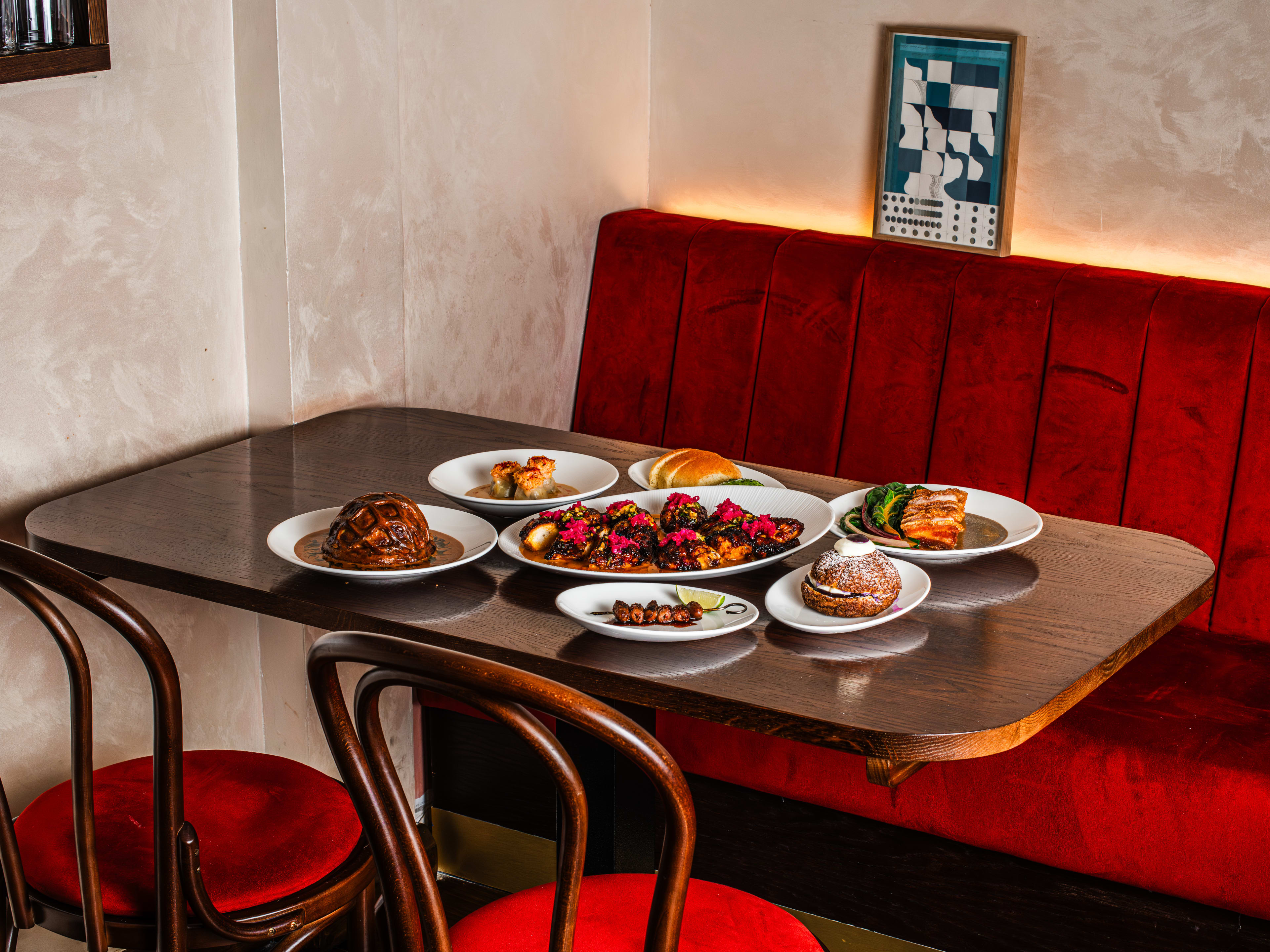 A group of dishes from Donia served on white plates sitting on a dark wooden table. There are Two cabaret chairs with red velvet cushions opposite  a red velvet booth.