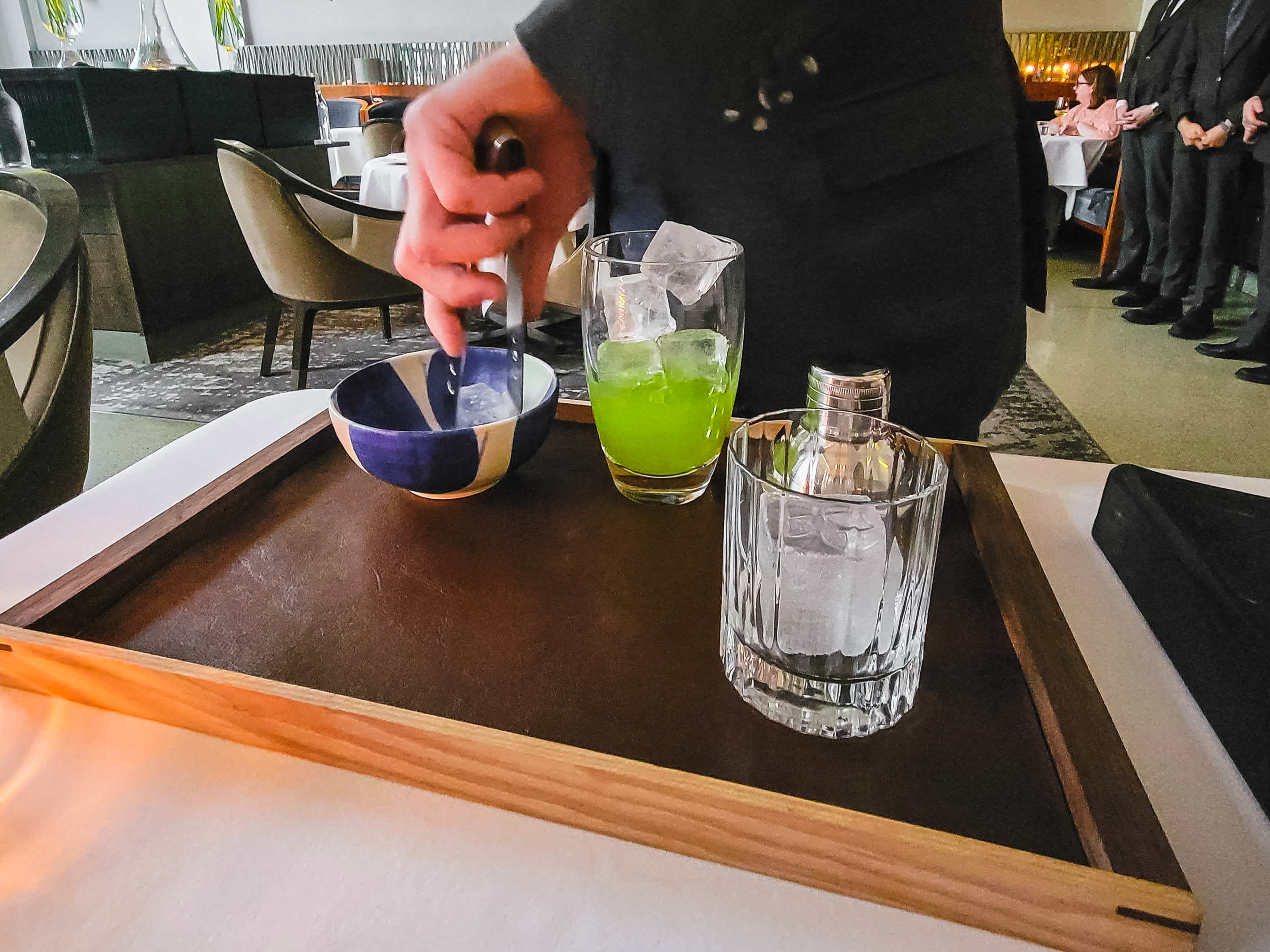 A server in a suit plucking ice cubes from a bowl with tongs and putting them into a glass of bright green juice.