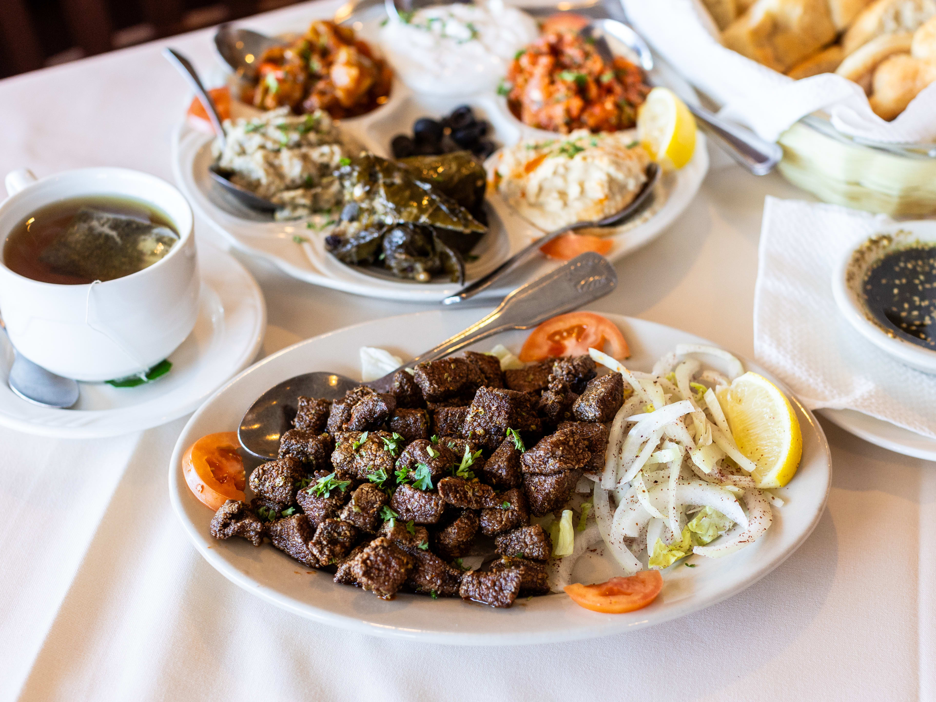 A plate of fried liver next to a cup of tea, a small bowl of sauce, and a mix platter and bread in the background.