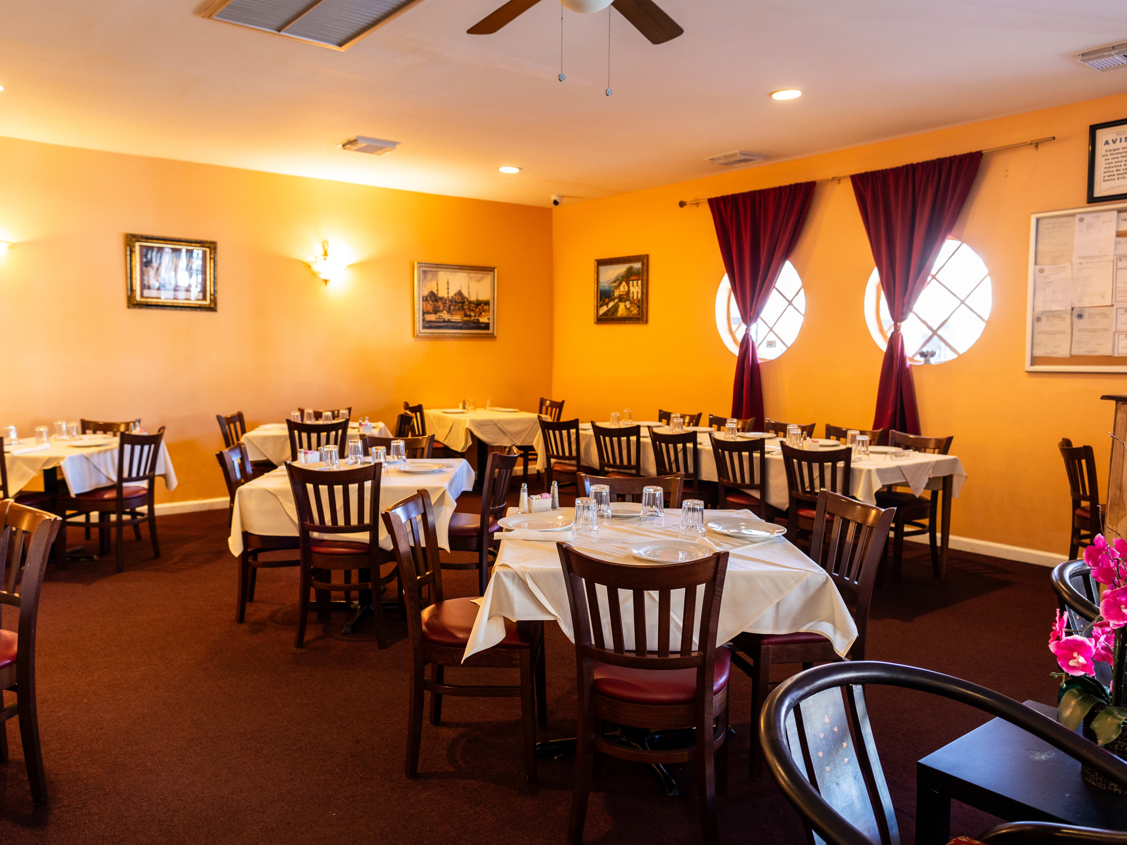 The interior of Empire Turkish Grill. With tables with white tablecloths and maroon accents in the carpet, chairs, and curtains.