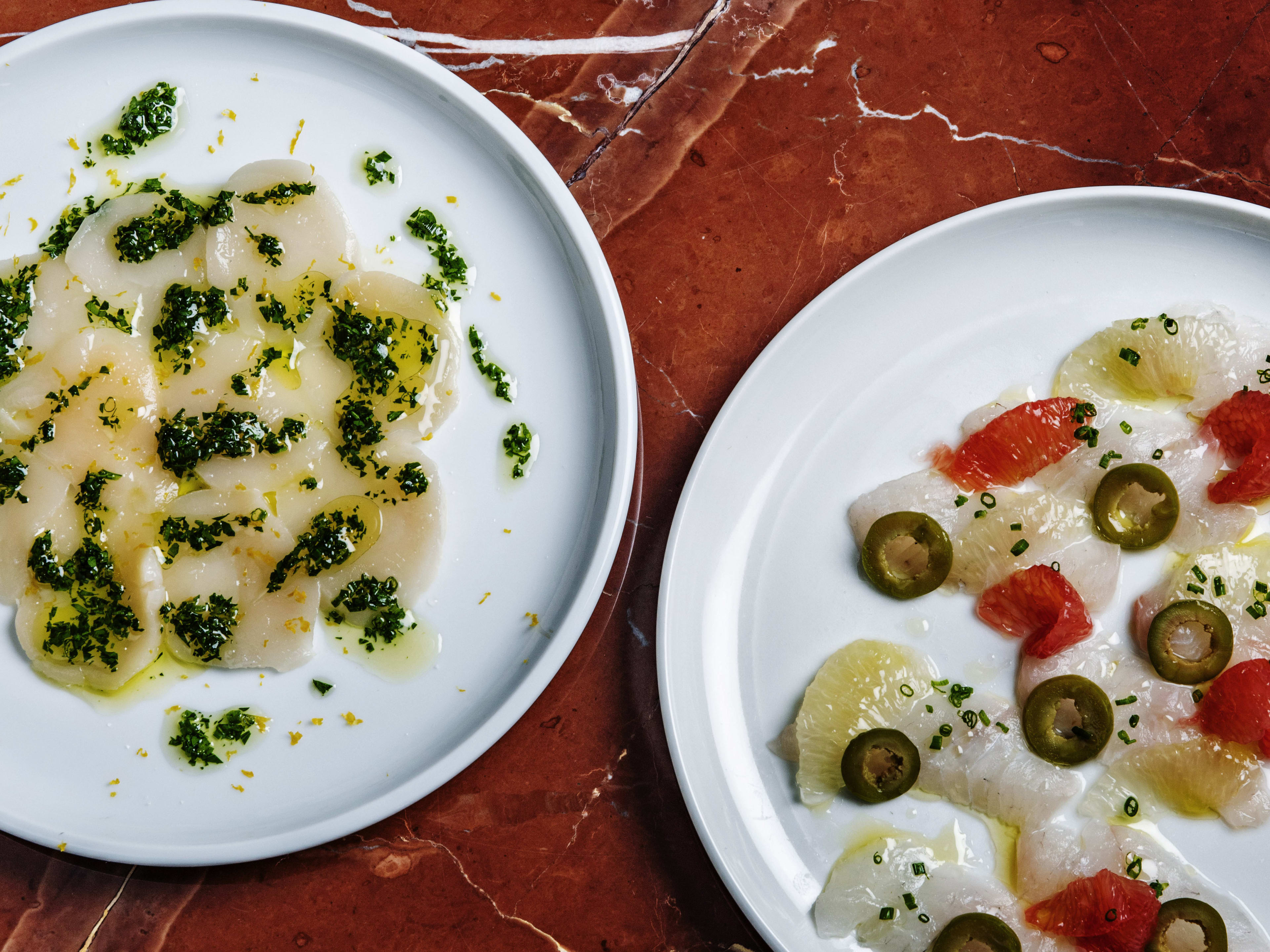 A plate of scallop crudo and a plate of fluke crudo on a red marble surface.