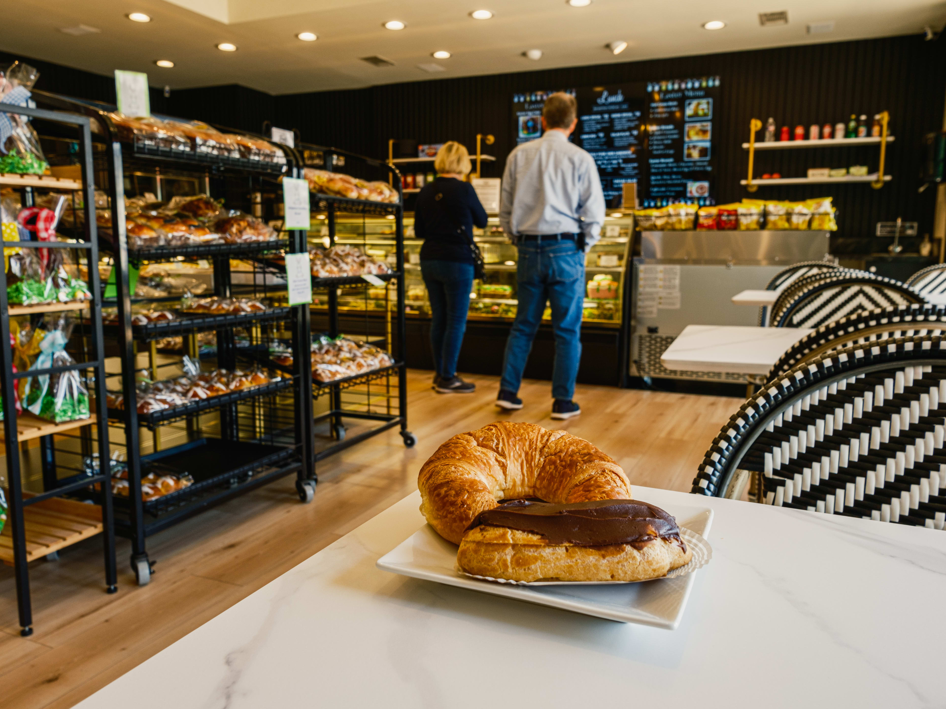 A chocolate eclair and a croissant on a table. Two people browse the pastry case at French Gourmet.