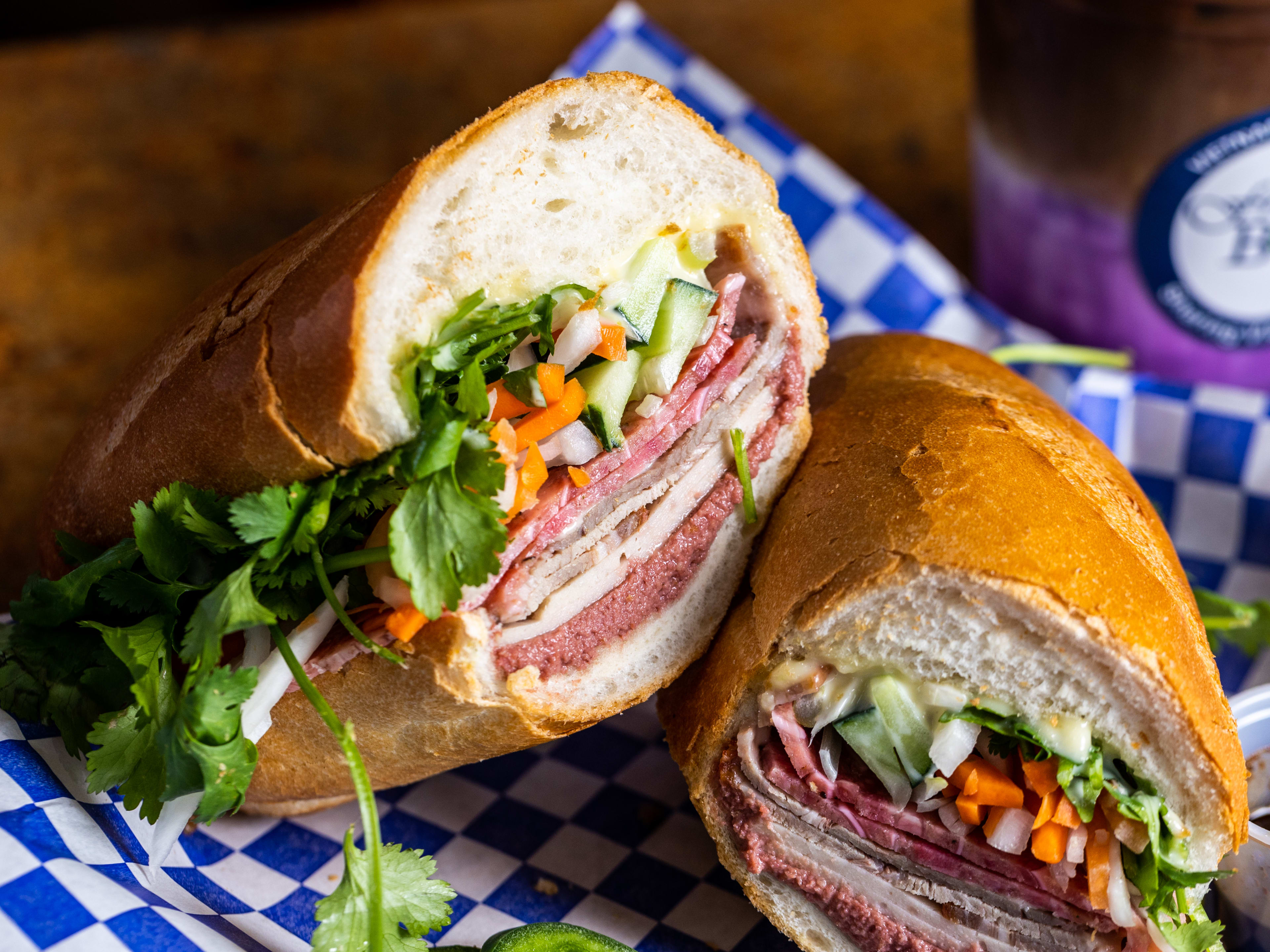 The house special banh mi from Fresh Bowl served cut in half on blue and white checkered paper.