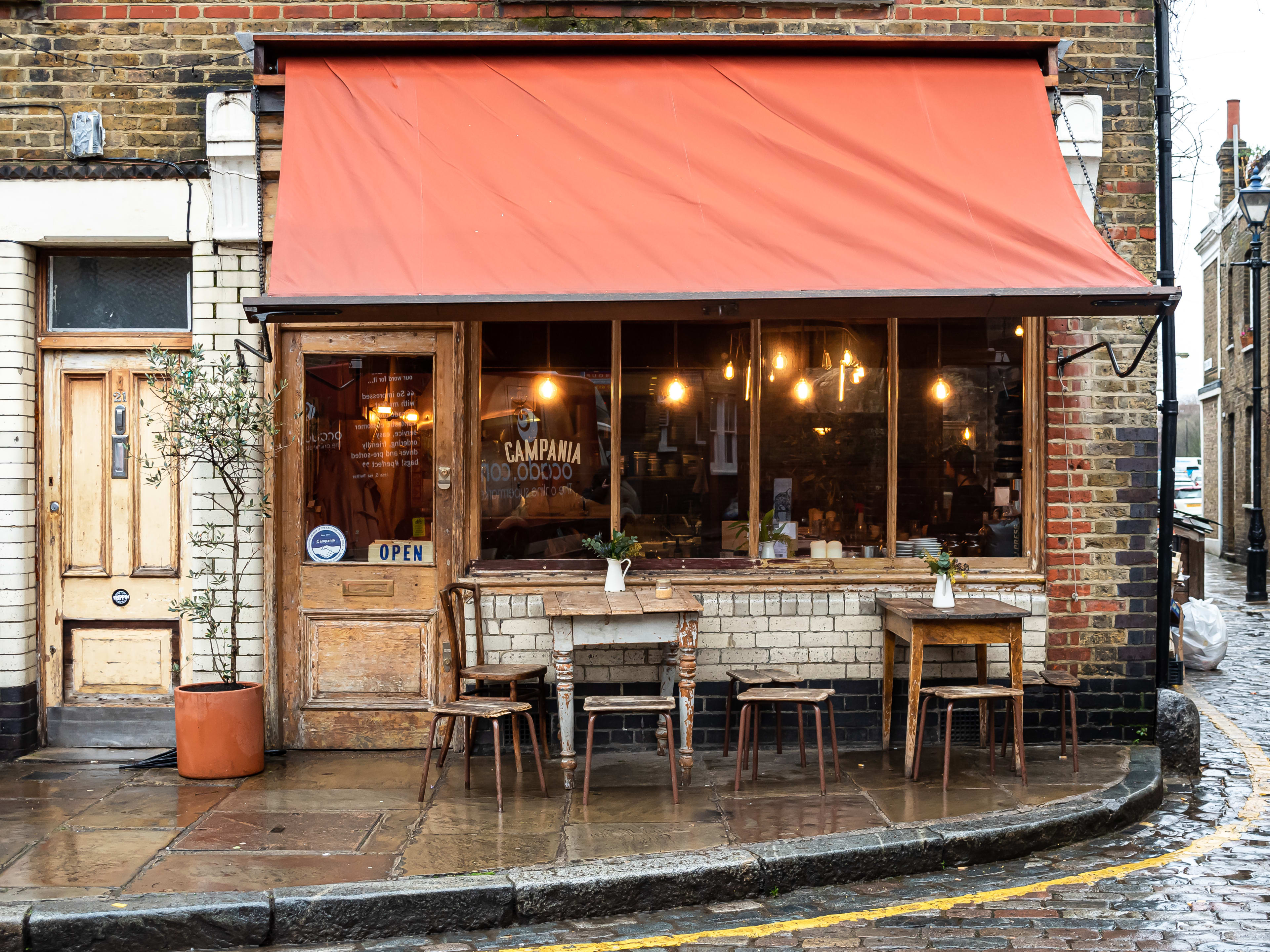 The brick exterior of Campania & Jones. The street is cobbleston, there are small wooden tables out front and warm lighting seen through the windows.