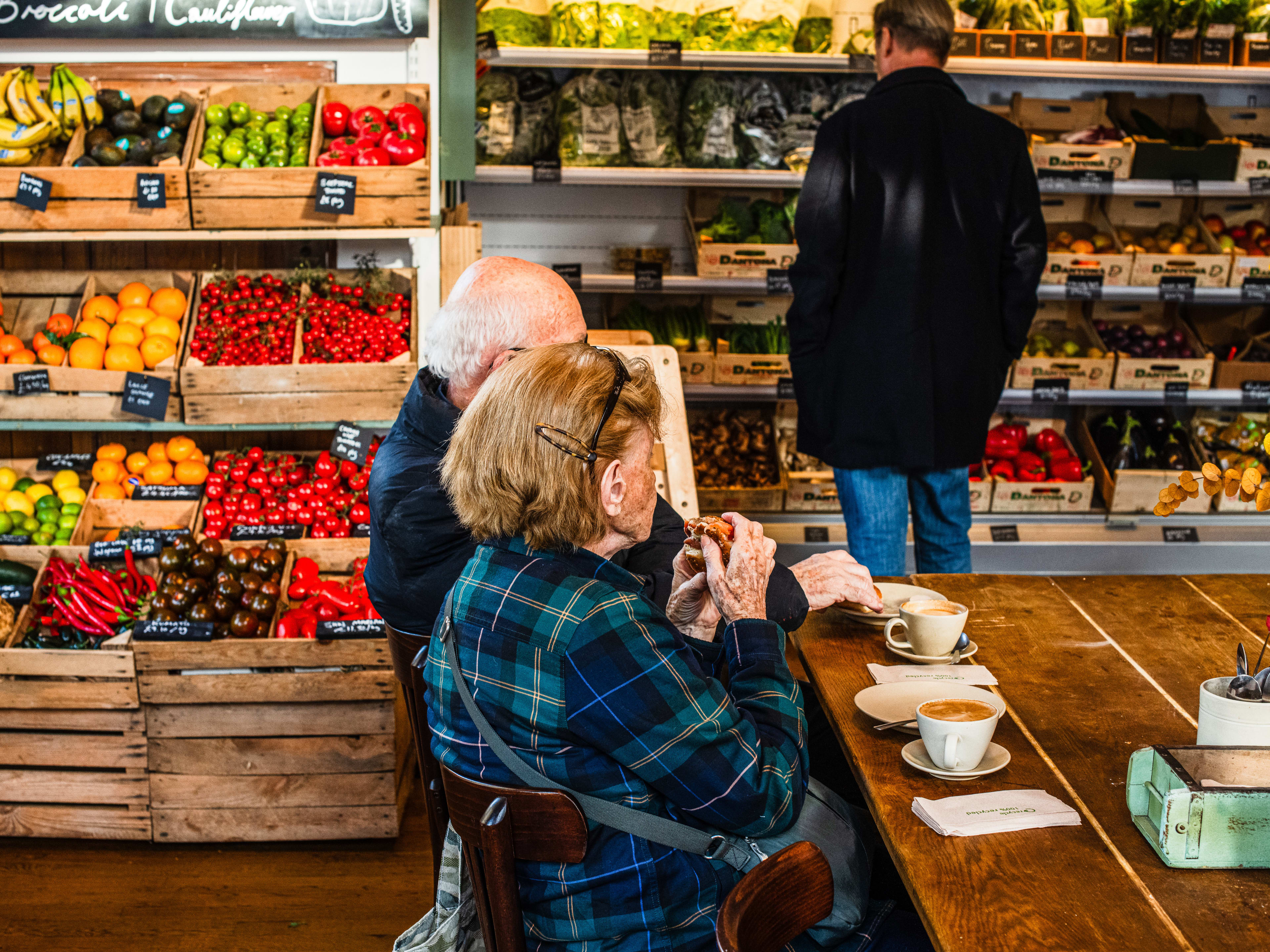 Two people sit at a table drinking coffee and eating pastries. There is a wall of produce behind them with someone browsing the items.